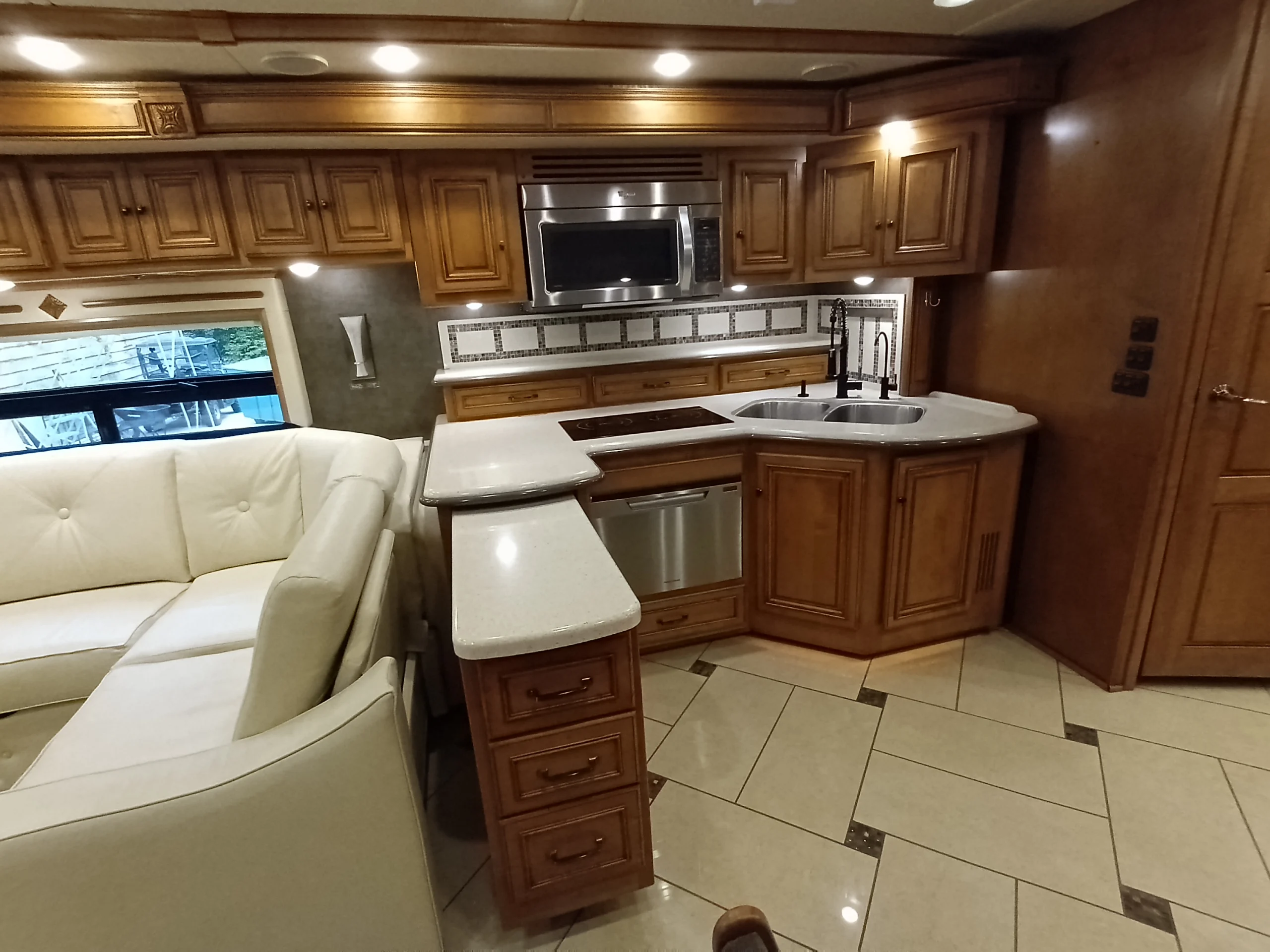 IMG_20260710_175114_599 Luxurious RV kitchen with wood cabinetry, polished countertops, built-in microwave, and corner sink. Cream sofa visible, elegant motorcoach design.