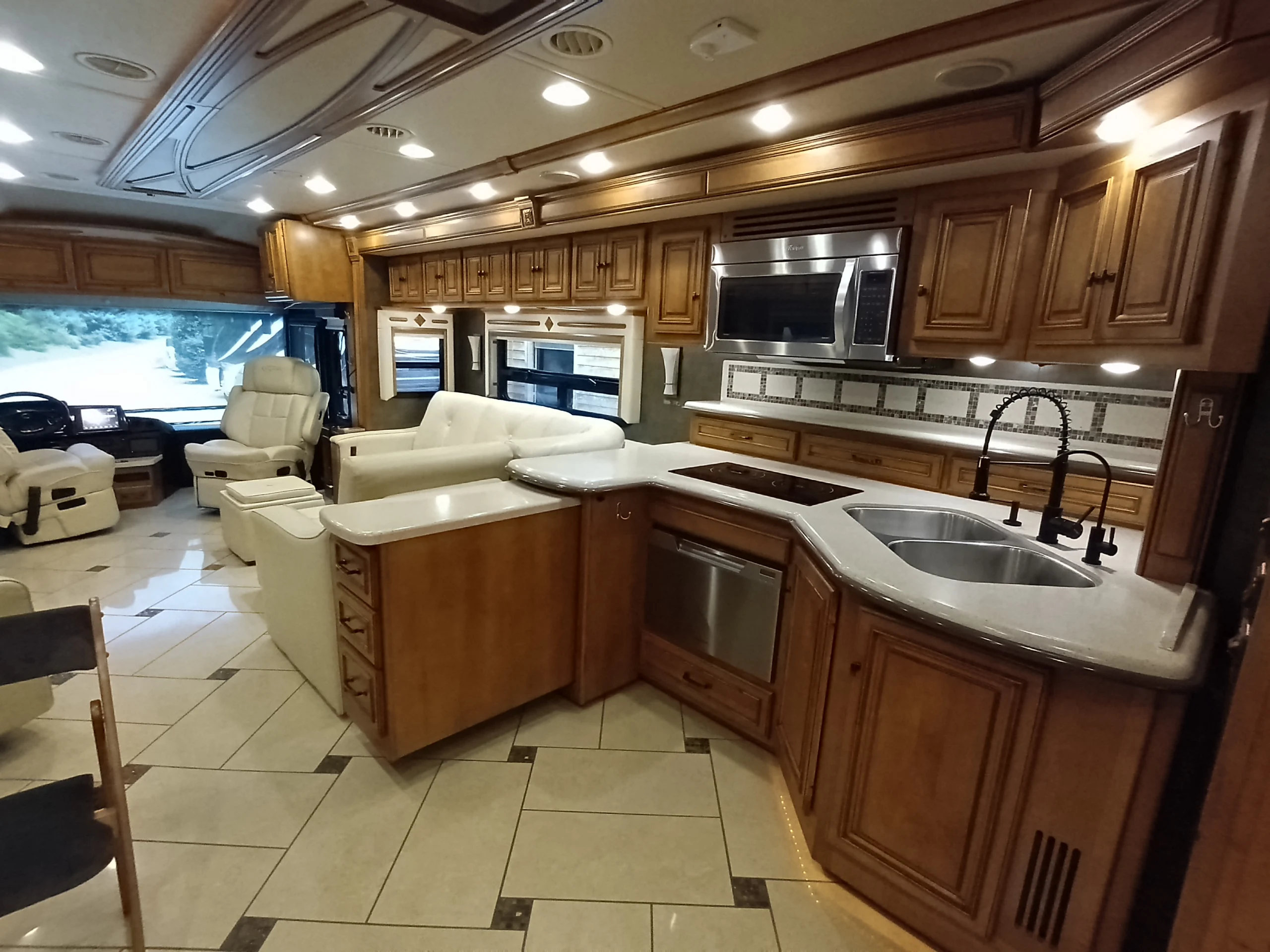 IMG_20260710_175126_350 Luxurious motorcoach interior featuring wooden cabinetry, white seating, a modern kitchen with stainless steel appliances, and tiled flooring.