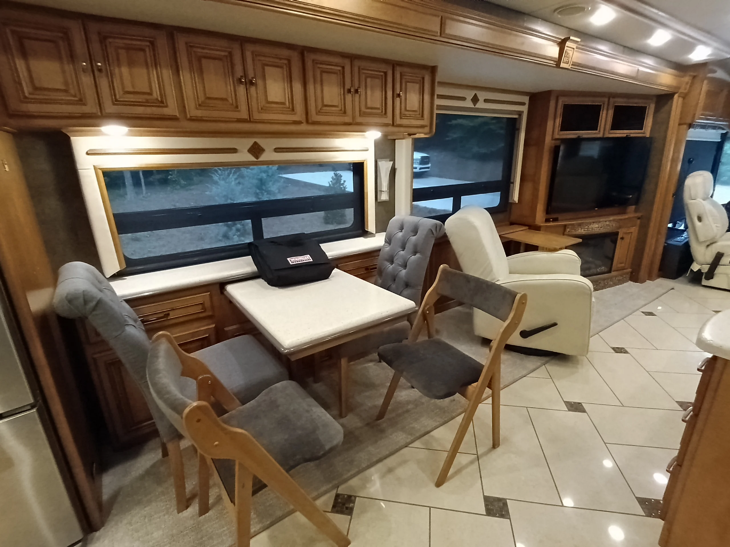 IMG_20260710_175135_750 Spacious Motorcoach RV interior with dining area, plush chairs, wooden cabinets, tiled floor, and large windows offering an outdoor view.