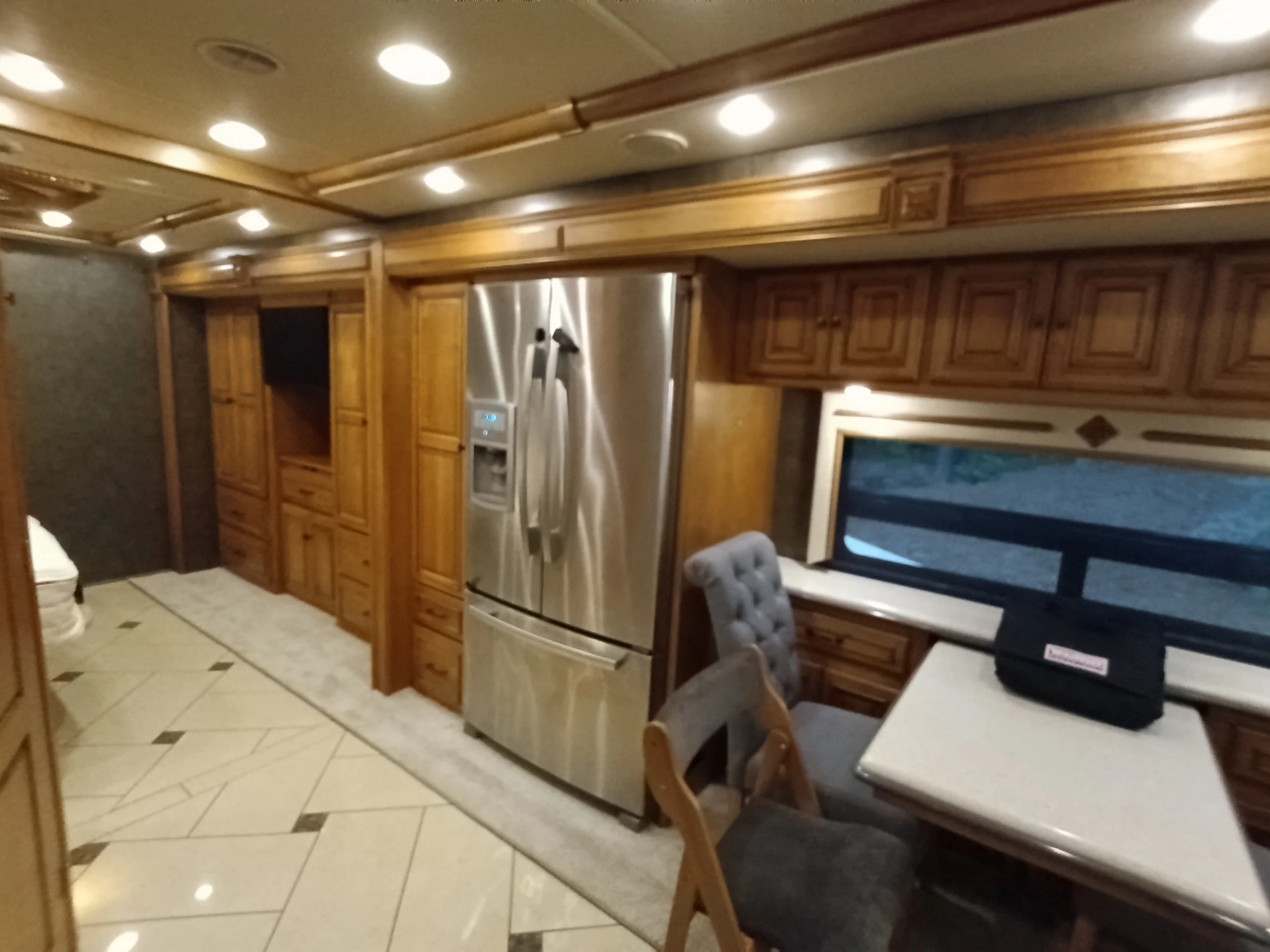IMG_20260710_175154_503 Luxurious Motorcoach interior featuring wooden cabinets, stainless steel fridge, tiled floor, and dining area with cushioned chairs.