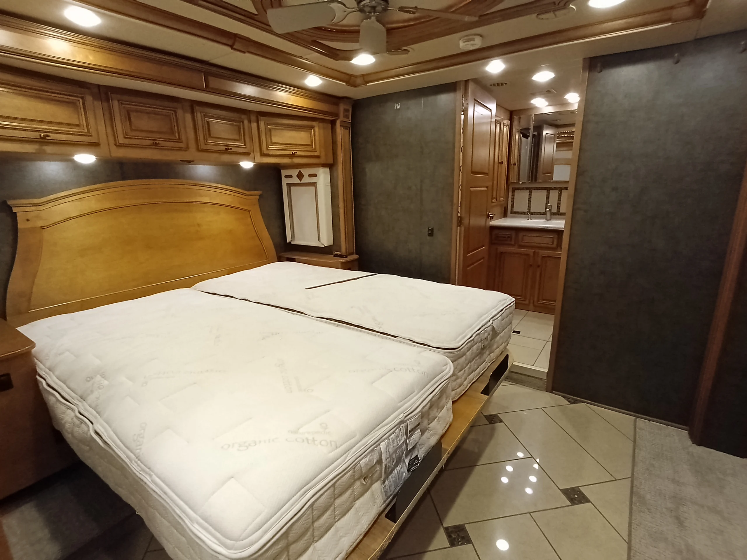 IMG_20260710_175213_352 Luxury motorcoach bedroom with twin beds, wooden cabinets, and en-suite bathroom. Comfortable, spacious interior for RV living.