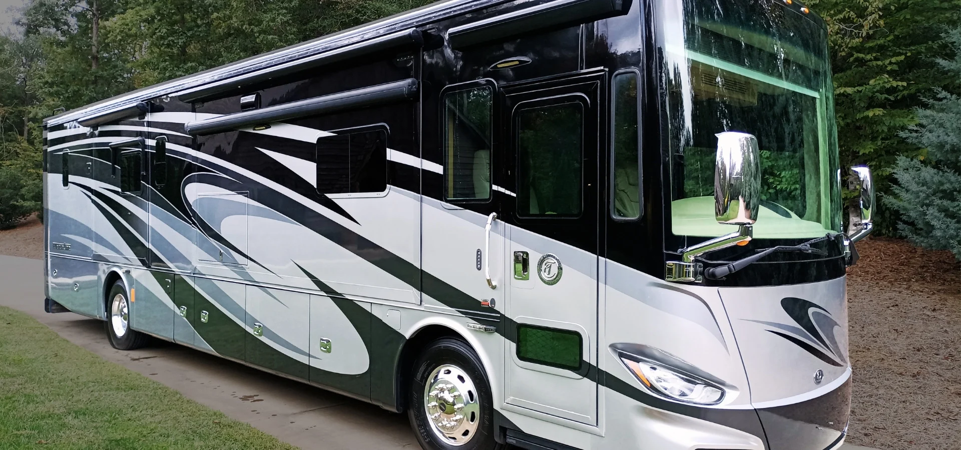 Luxury motorcoach with sleek black and silver design parked on a driveway, surrounded by trees.