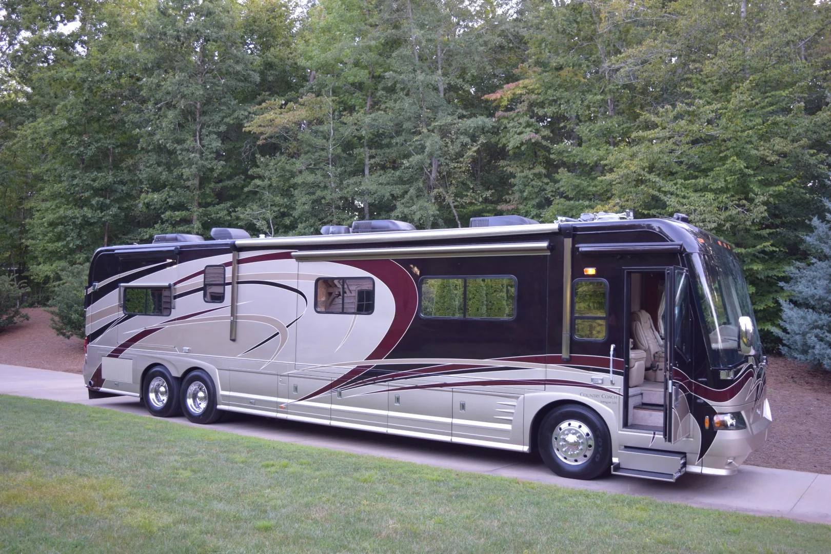 Large, luxurious Motorcoach parked on driveway, surrounded by lush greenery, featuring sleek design with maroon and beige accents.