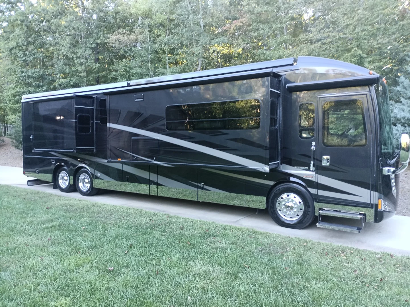 Sleek black motorcoach with extended slides parked on a driveway, surrounded by green trees and grass.