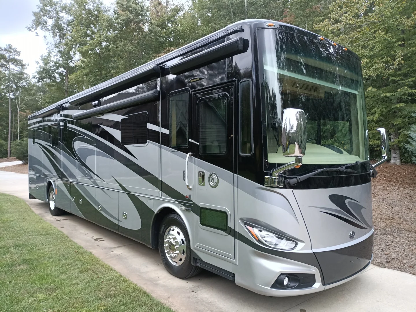 Sleek, modern Motorcoach and RV parked on a driveway amidst greenery, featuring a black and gray exterior with large windows.