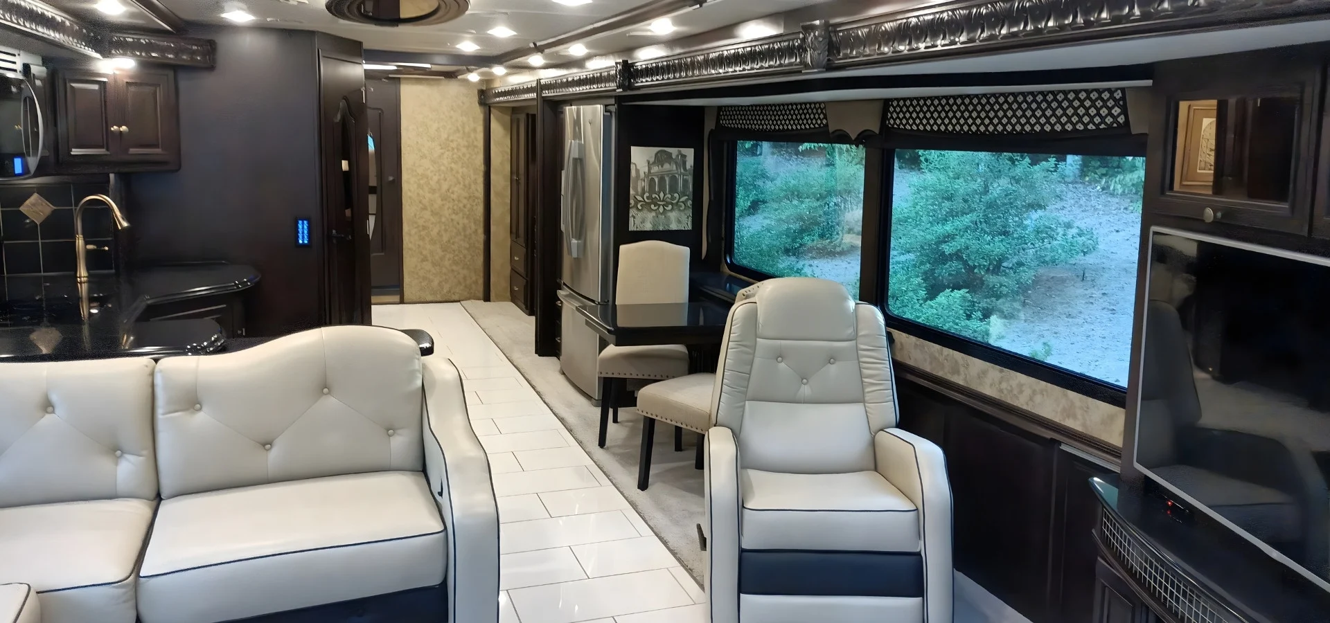 Luxurious motorcoach interior with beige leather seating, dining area, stainless steel appliances, and large windows displaying greenery.