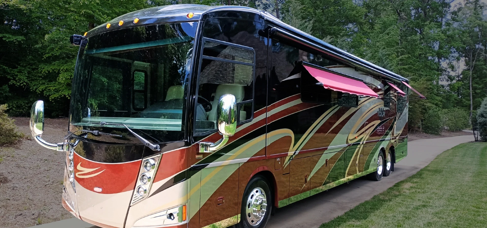 Luxurious motorcoach with colorful swirls, reflective side mirrors, and red window awnings parked on a tree-lined road.