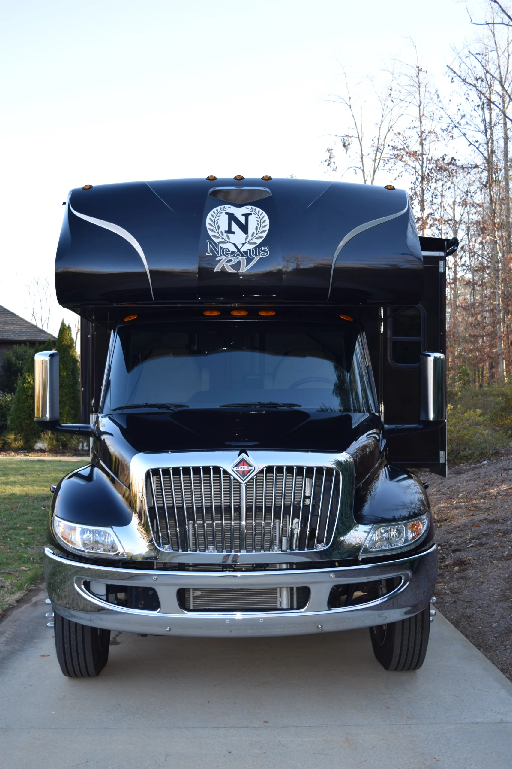 DSC_0002 Black Nexus RV motorcoach parked on a driveway, surrounded by trees, under a clear blue sky.