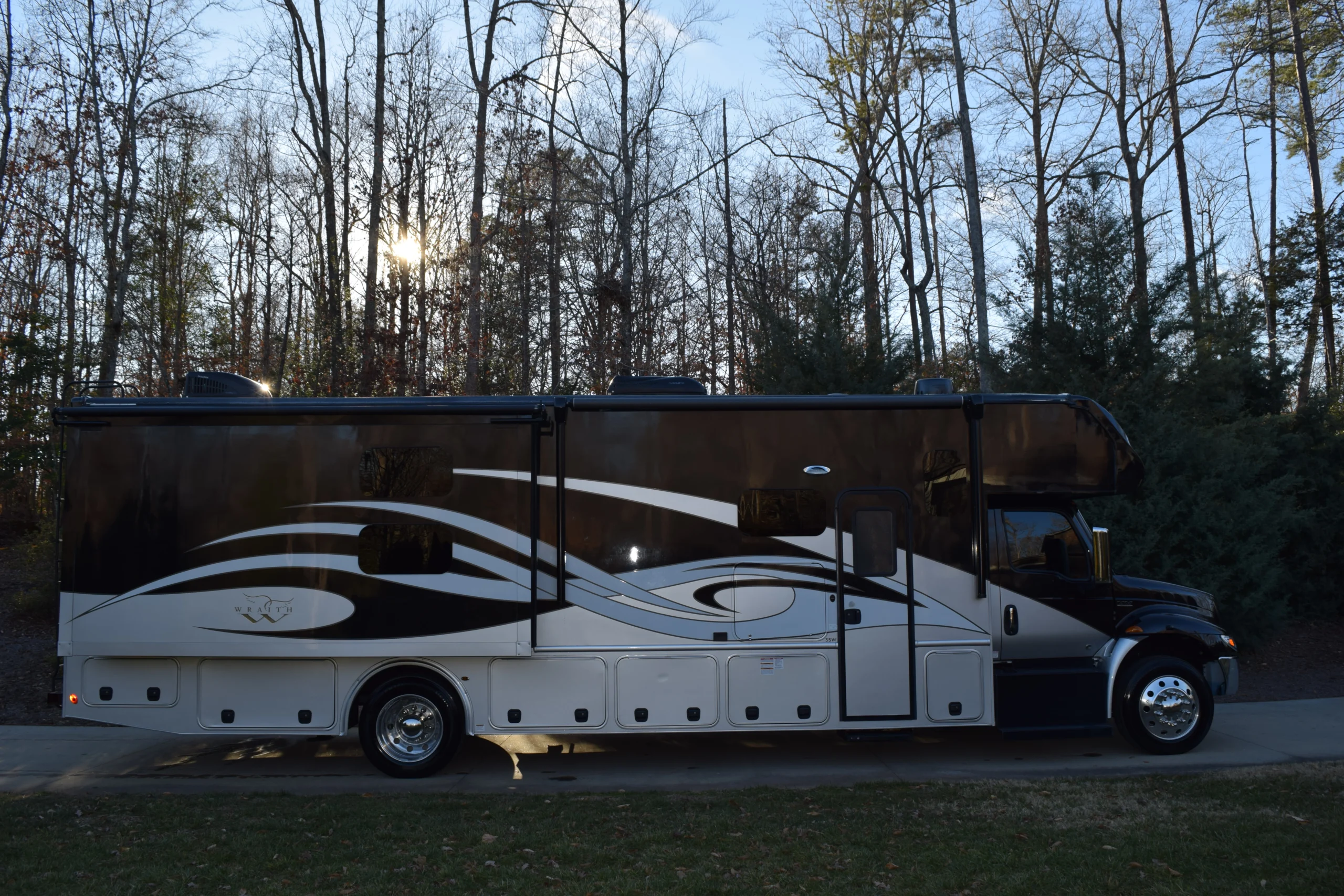 DSC_0009 Luxurious motorcoach with sleek design parked on a driveway, surrounded by tall trees and a setting sun in the background.