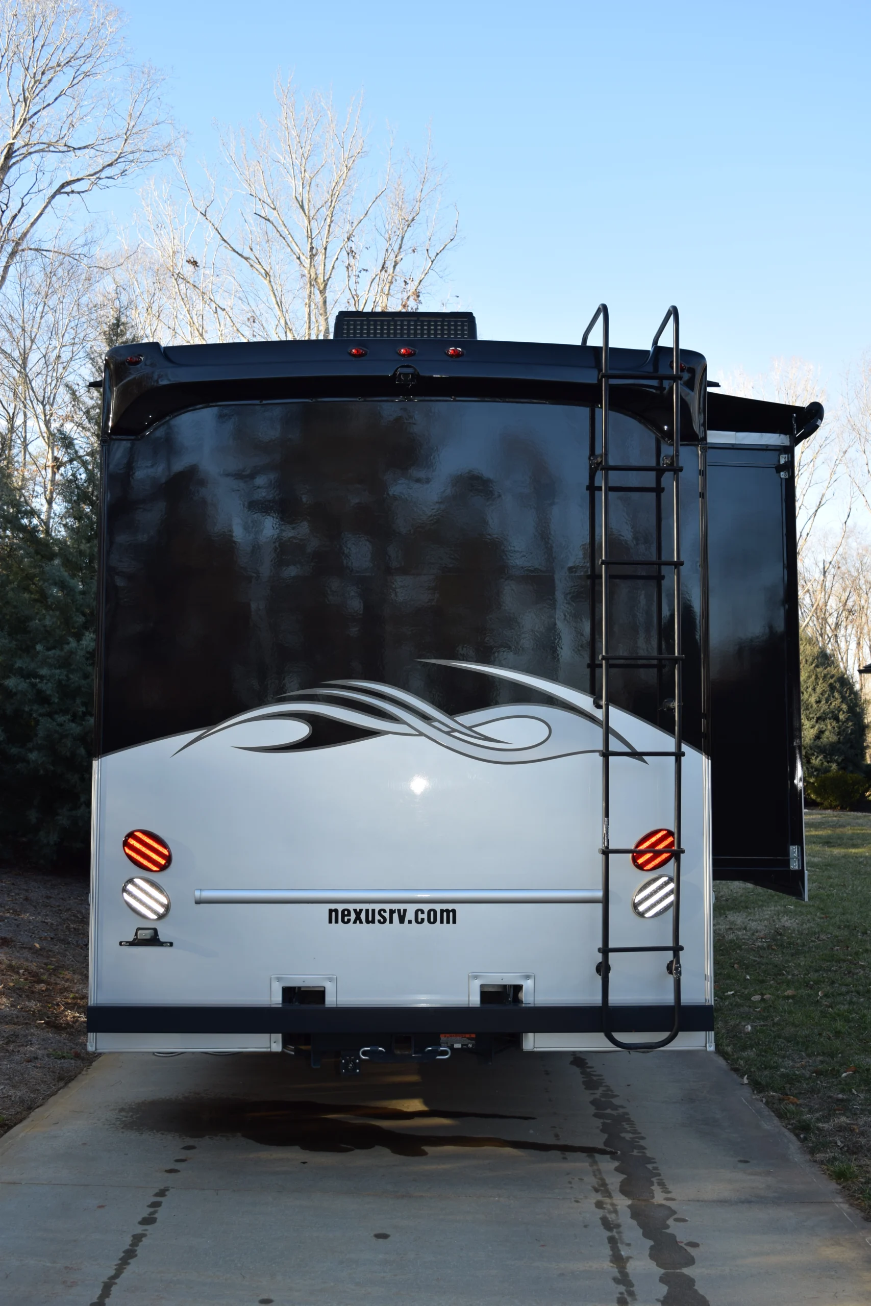 DSC_0010 Rear view of a motorcoach RV with a ladder and decorative swirl pattern, parked on concrete with trees in the background.