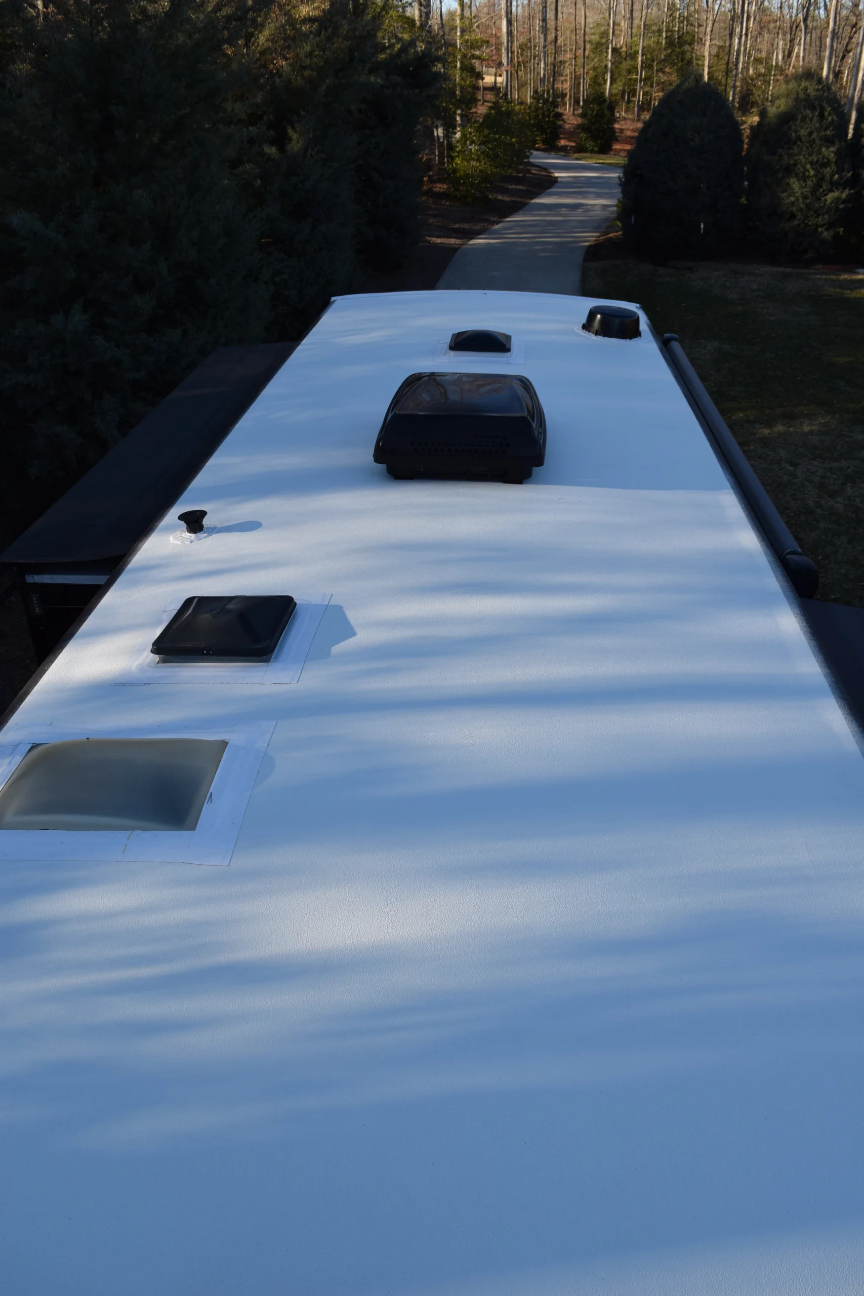 DSC_0011 Roof of a motorcoach with vents and skylights, surrounded by trees and a driveway in the background.