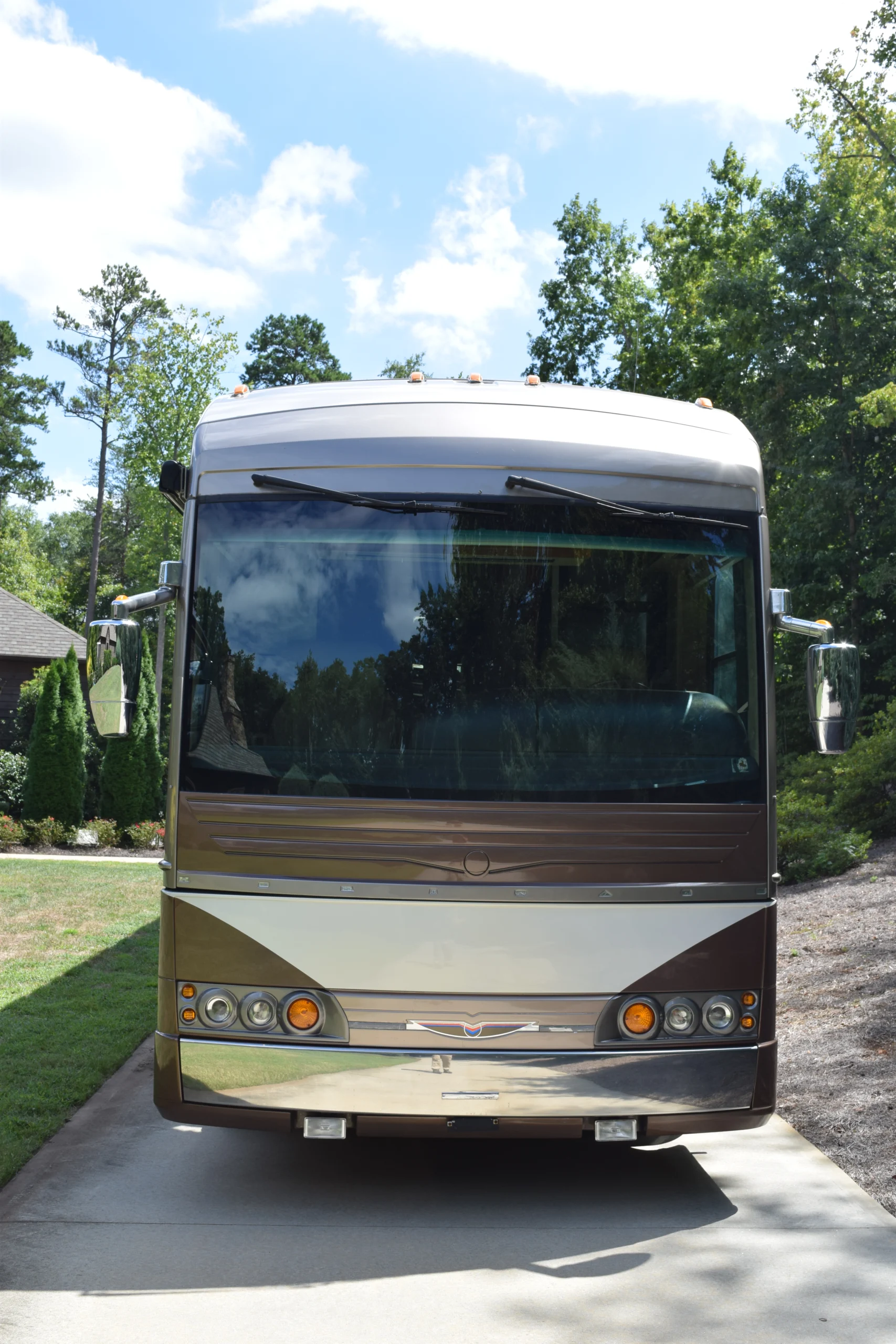 DSC_0036 Front view of a motorcoach parked on a driveway, surrounded by trees and a clear sky.