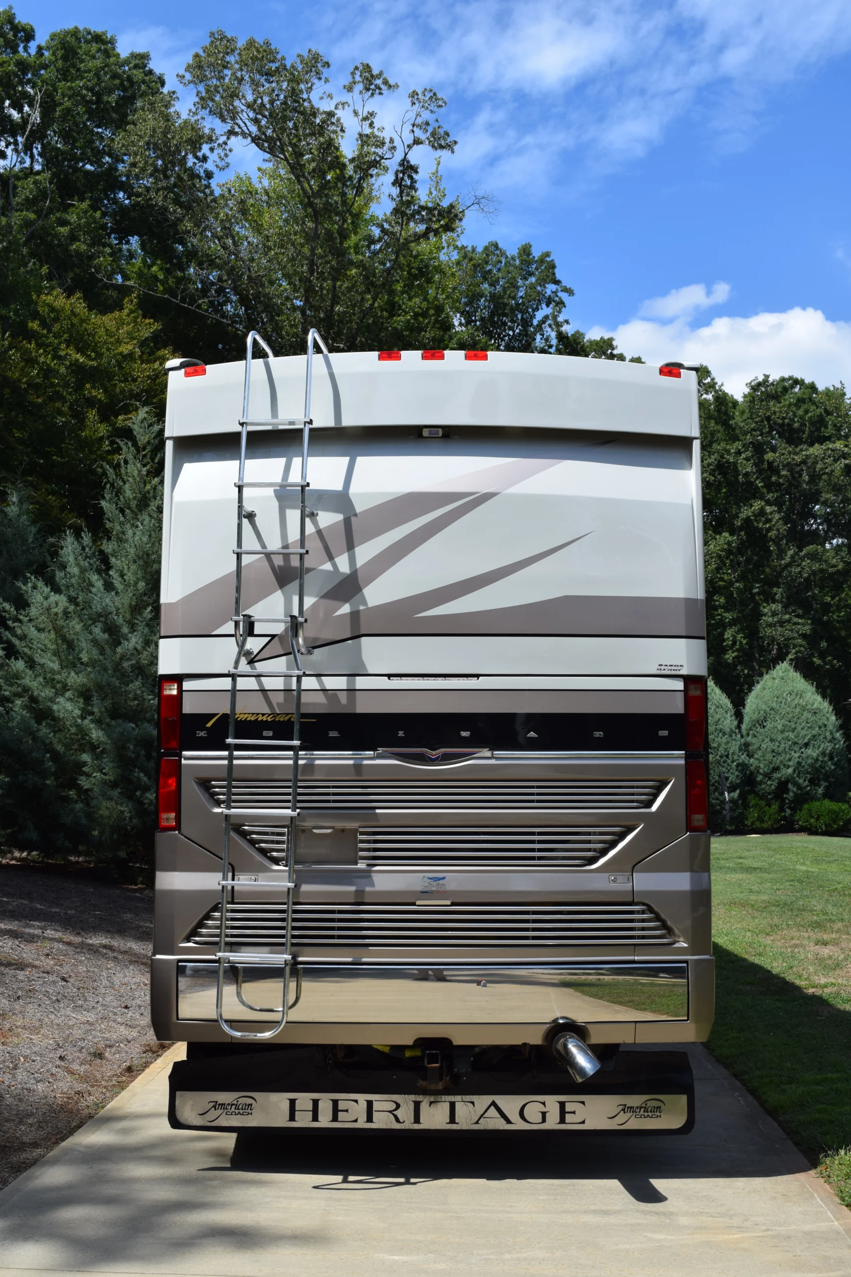DSC_0038 Rear view of a parked RV featuring a ladder, surrounded by trees under a clear blue sky.