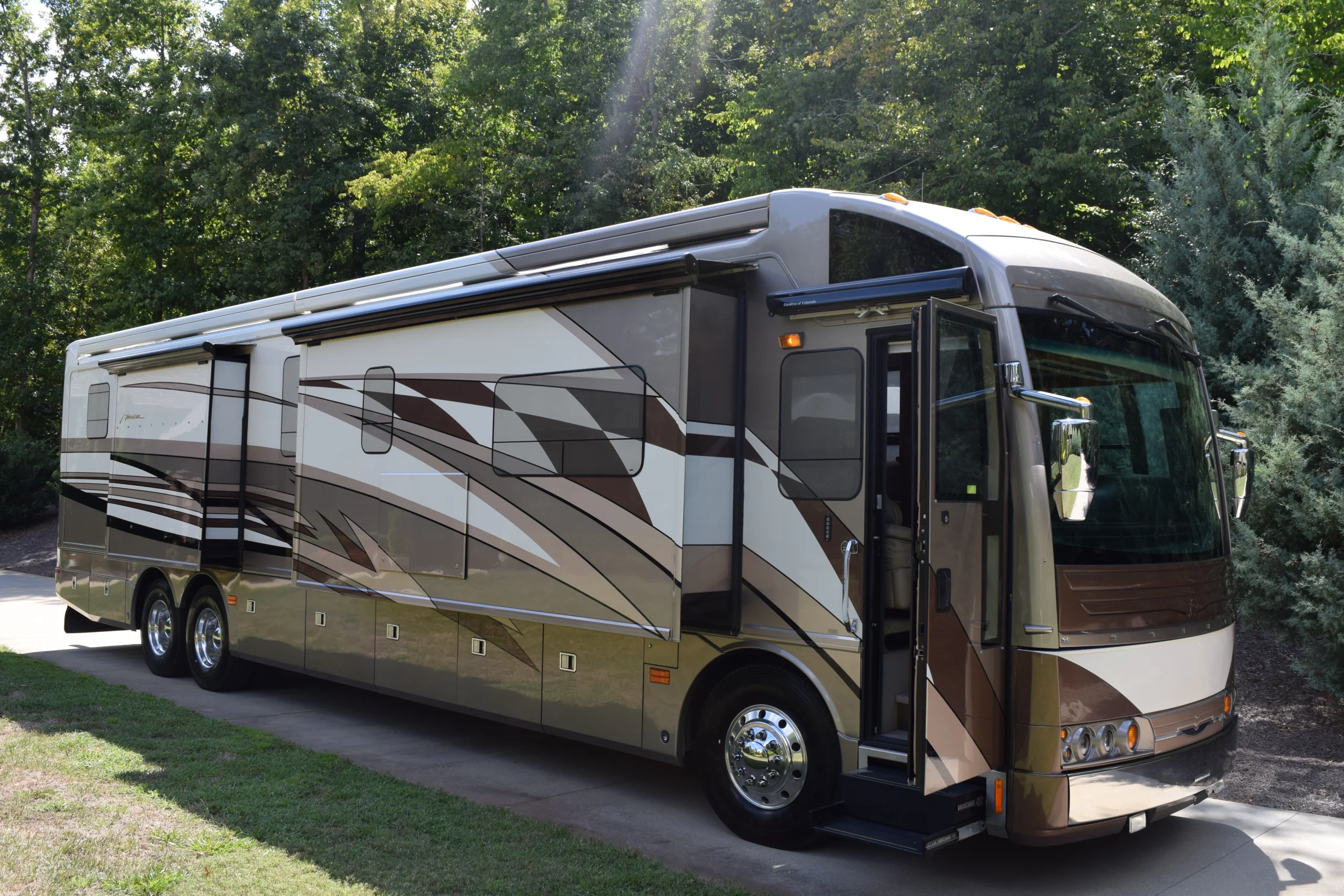 DSC_0040 Luxurious brown and beige motorcoach parked on a driveway, surrounded by lush trees and greenery.
