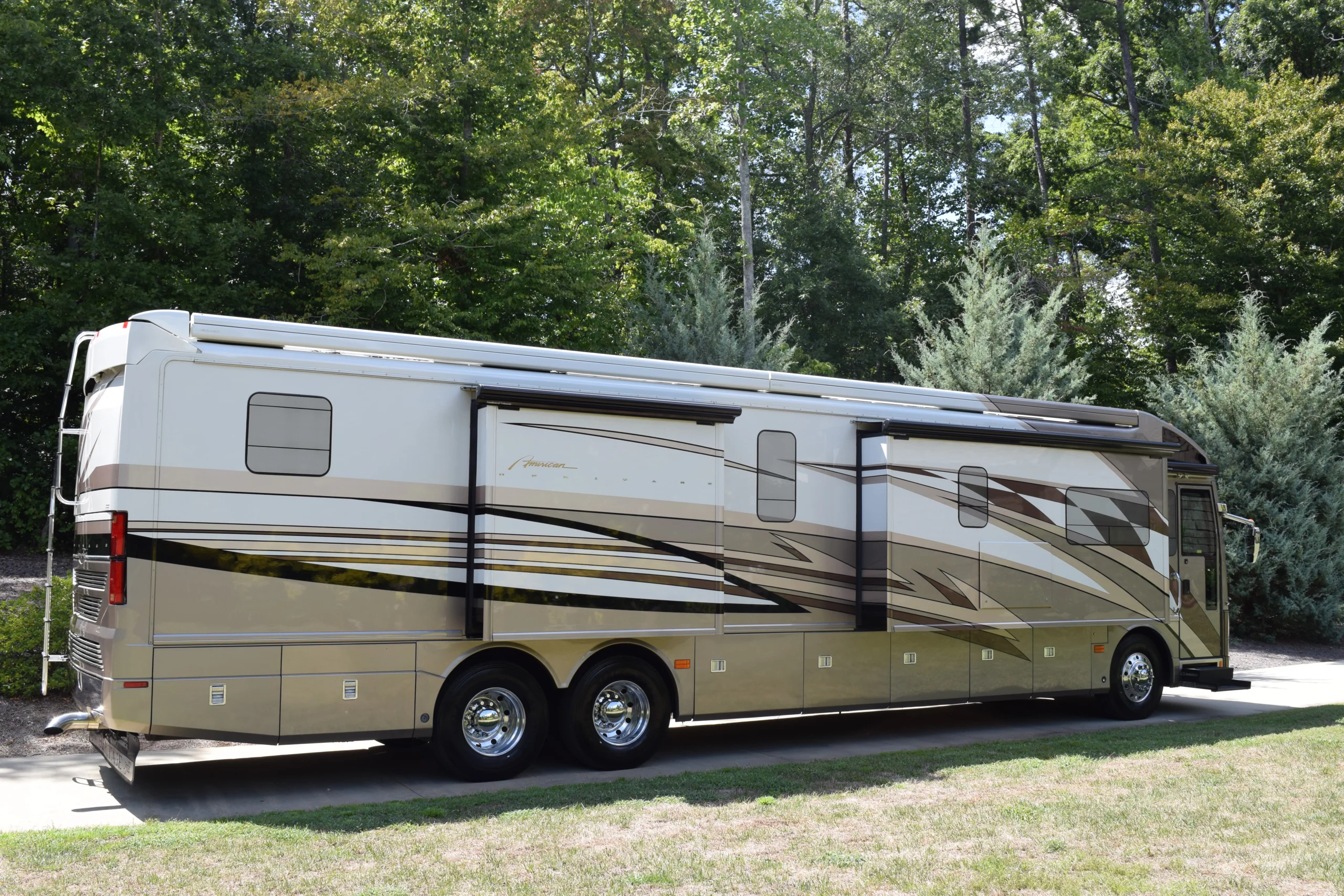 DSC_0042 Luxurious motorcoach with sleek beige and brown design, parked on a paved path, surrounded by lush green trees and grass.