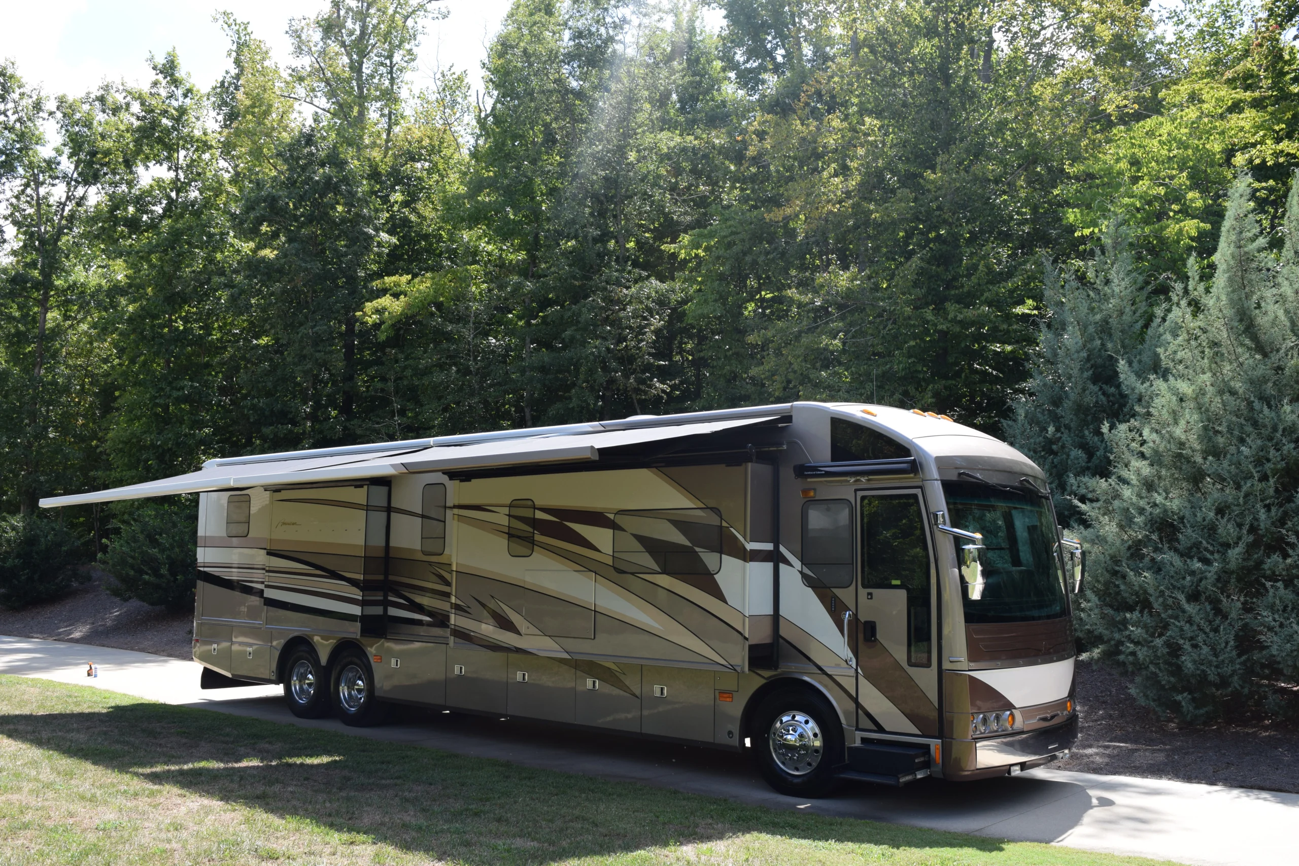DSC_0043 Large beige motorcoach RV with extended awning, parked on a driveway surrounded by lush trees.