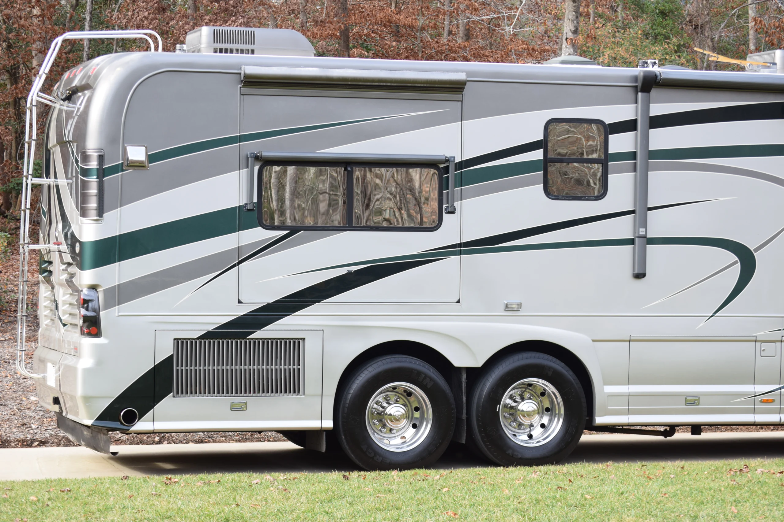 DSC_0138 Side view of a parked motorcoach with green and gray stripes, showcasing its dual rear wheels and attached ladder.