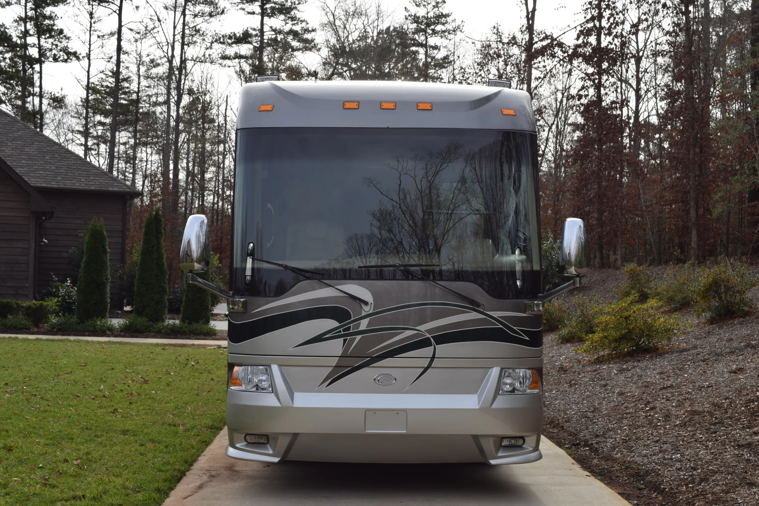 DSC_0142 Front view of a sleek motorcoach at the driveway, surrounded by trees and greenery.