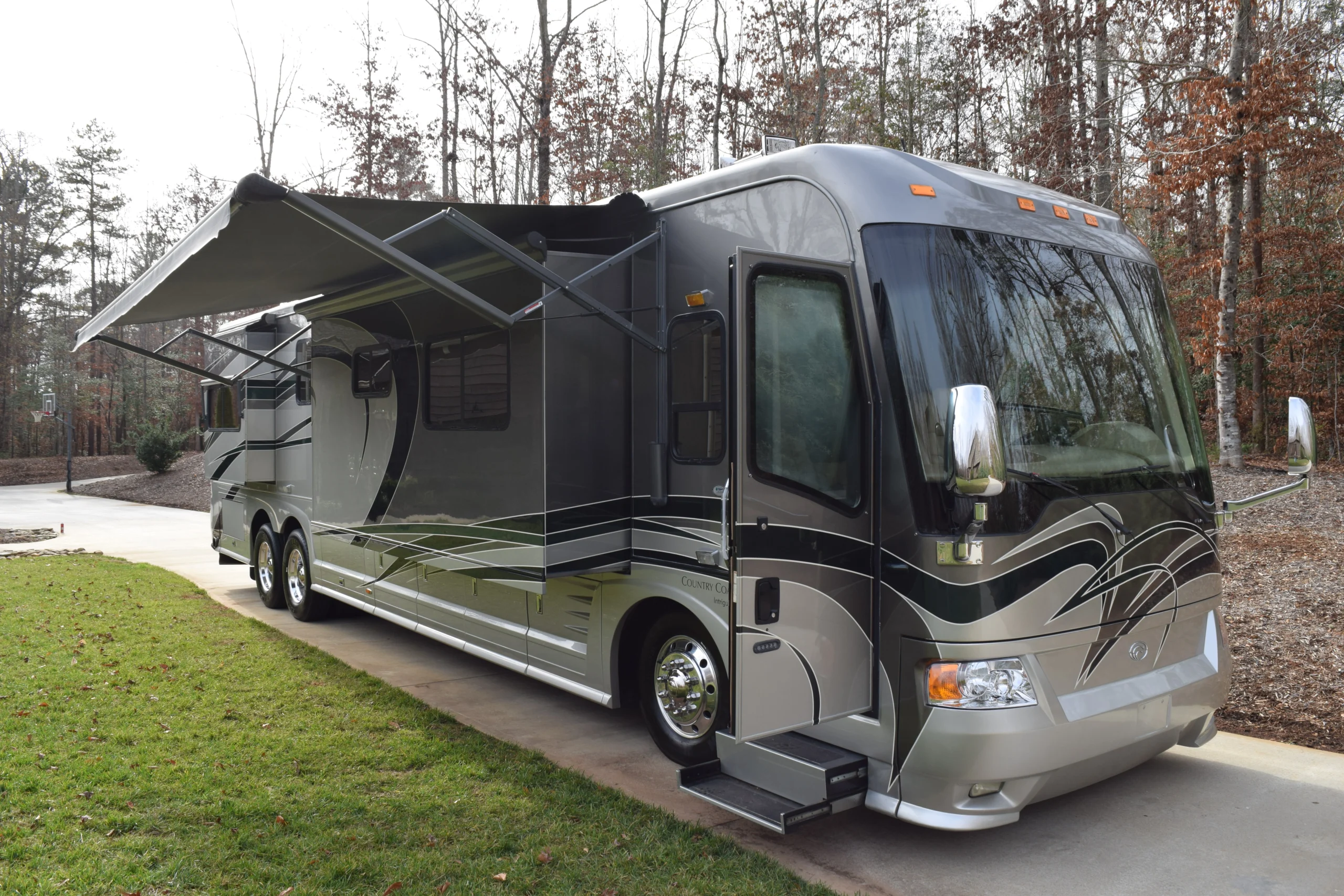 DSC_0148 Luxurious motorcoach with extended awnings parked on driveway, surrounded by autumn trees.