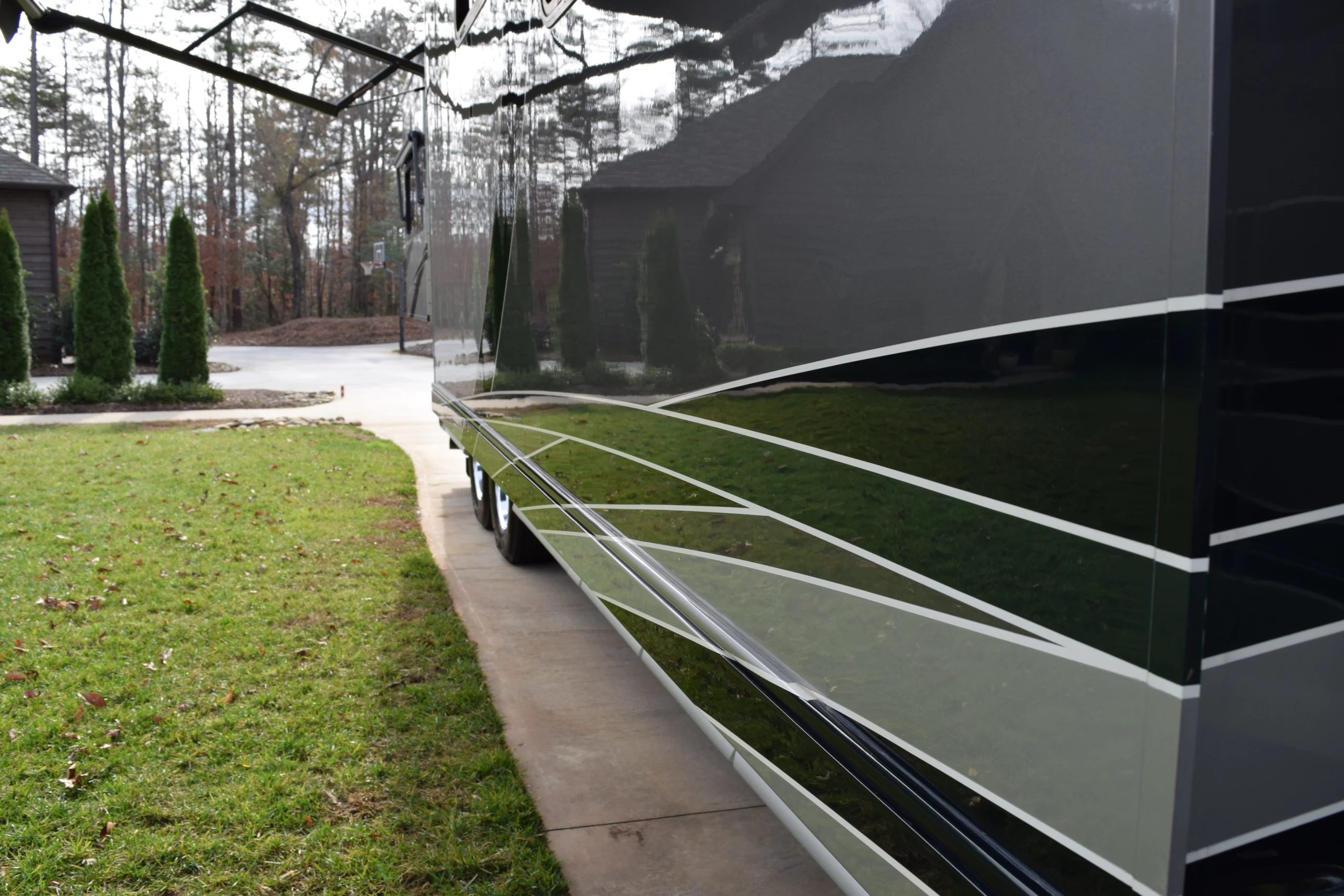 DSC_0156 Side view of a sleek, reflective motorcoach parked on a driveway, surrounded by greenery and trees in the background.