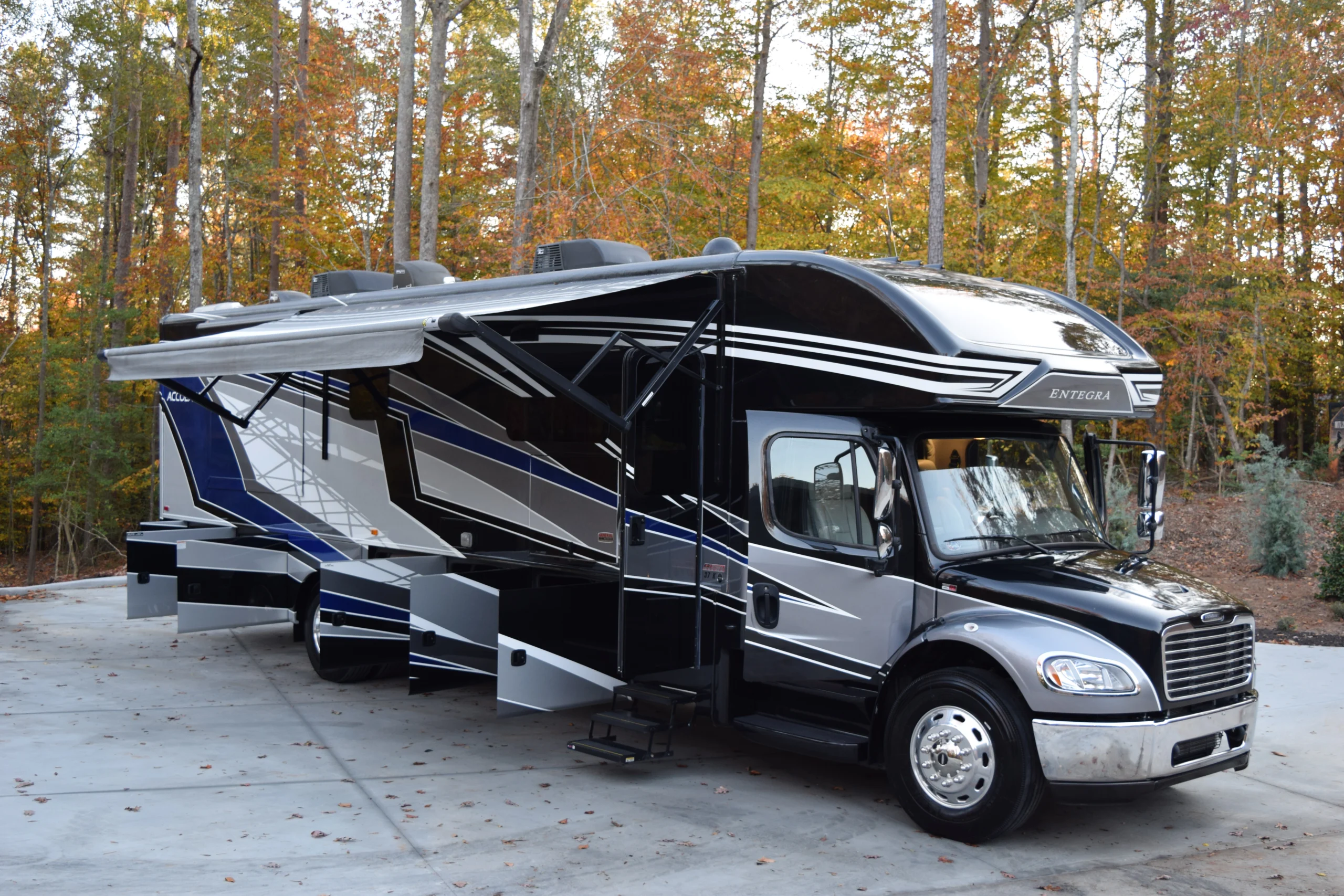 DSC_0171 Luxurious motorcoach with extended awning and slide-outs, parked on concrete surrounded by autumn trees.