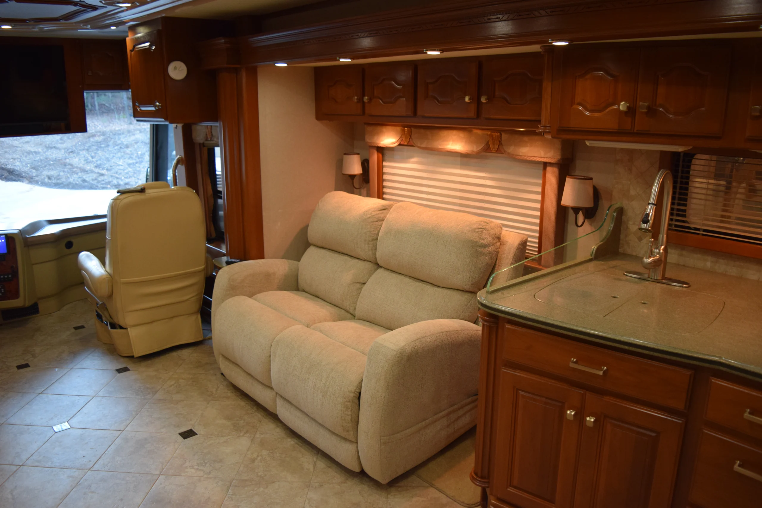 DSC_0173 Spacious motorcoach interior with beige sofa, recliner, and kitchenette. Luxurious wood cabinets and tiled floor enhance comfort and style.