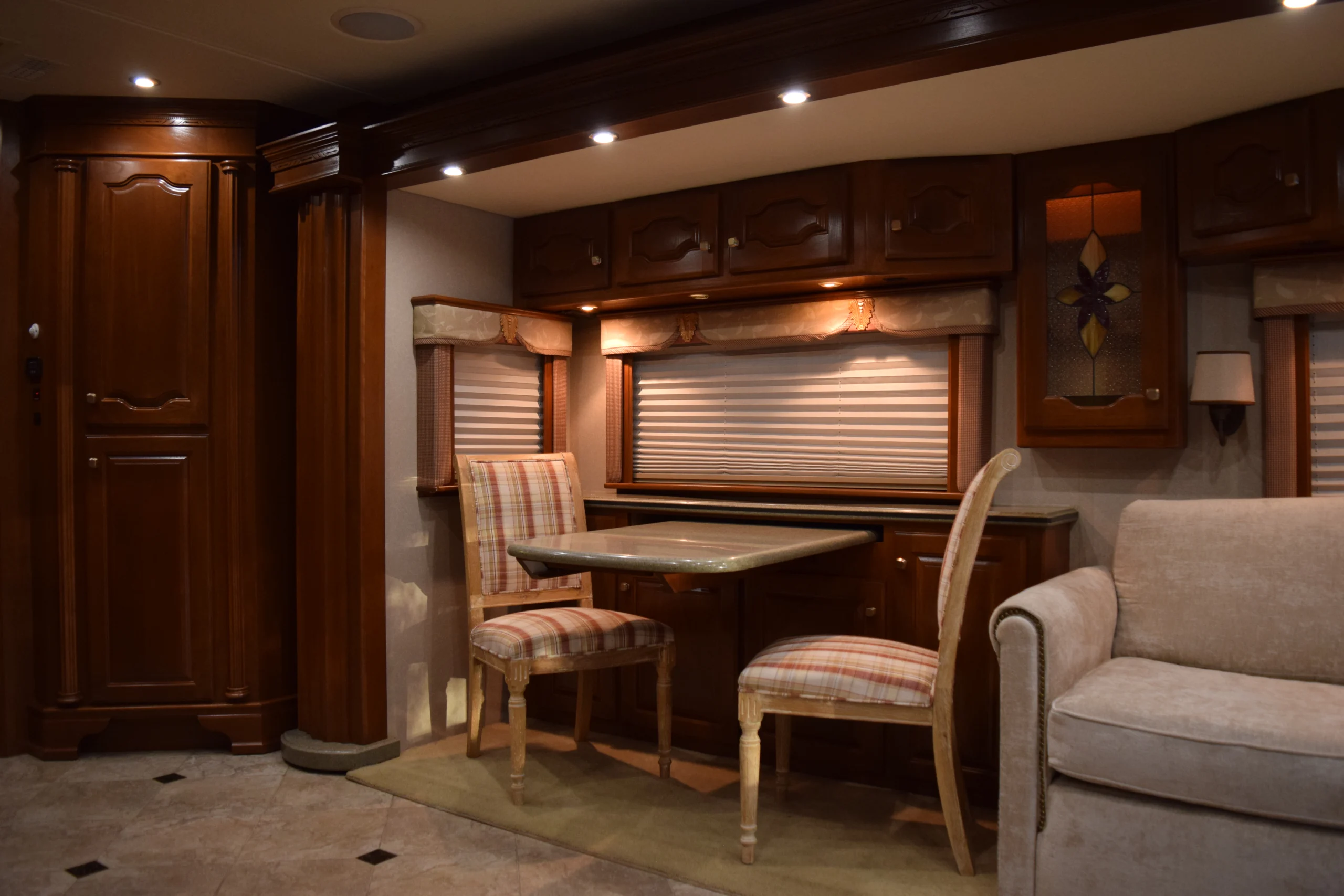 DSC_0187 Cozy Motorcoach interior featuring wooden cabinets, dining area with checkered chairs, and a beige sofa under warm lighting.