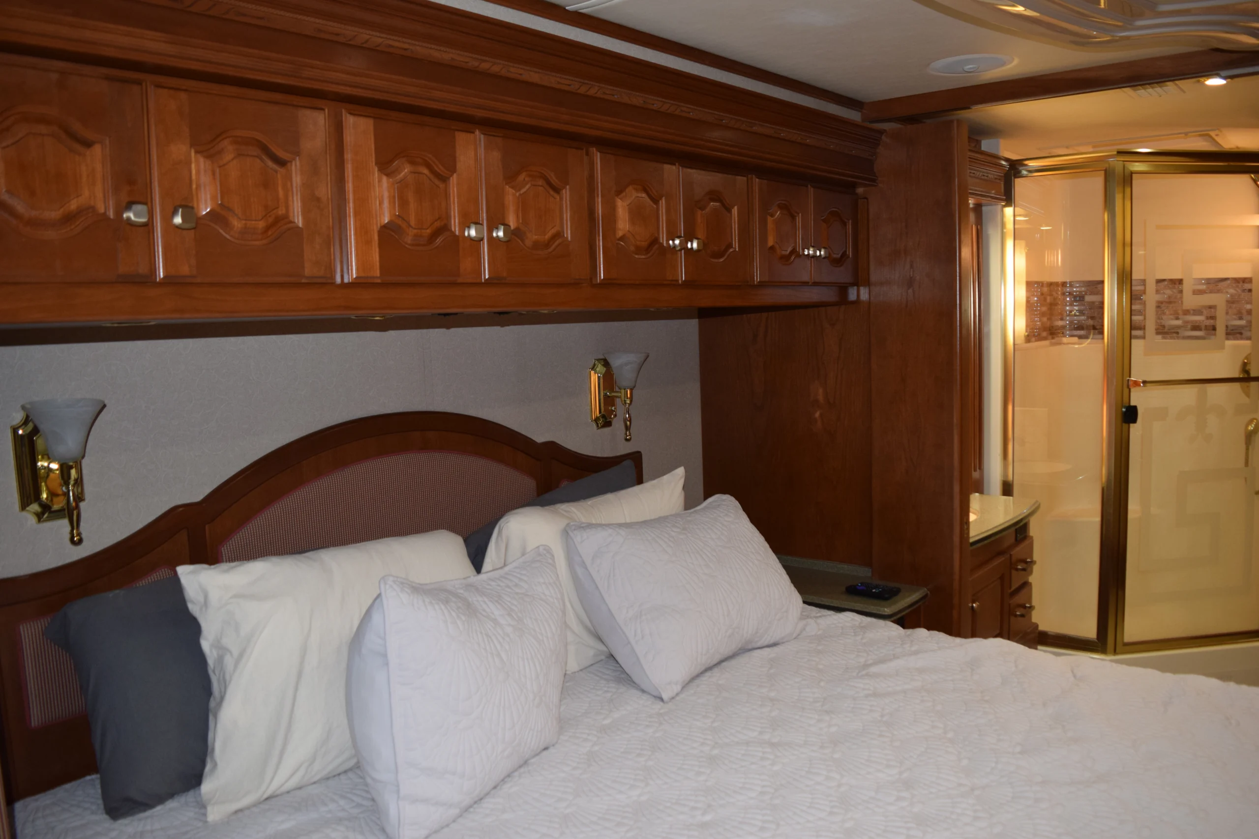 DSC_0192 Luxurious Motorcoach RV bedroom with wooden cabinetry, plush bed, white pillows, and an ensuite bathroom with glass shower door.