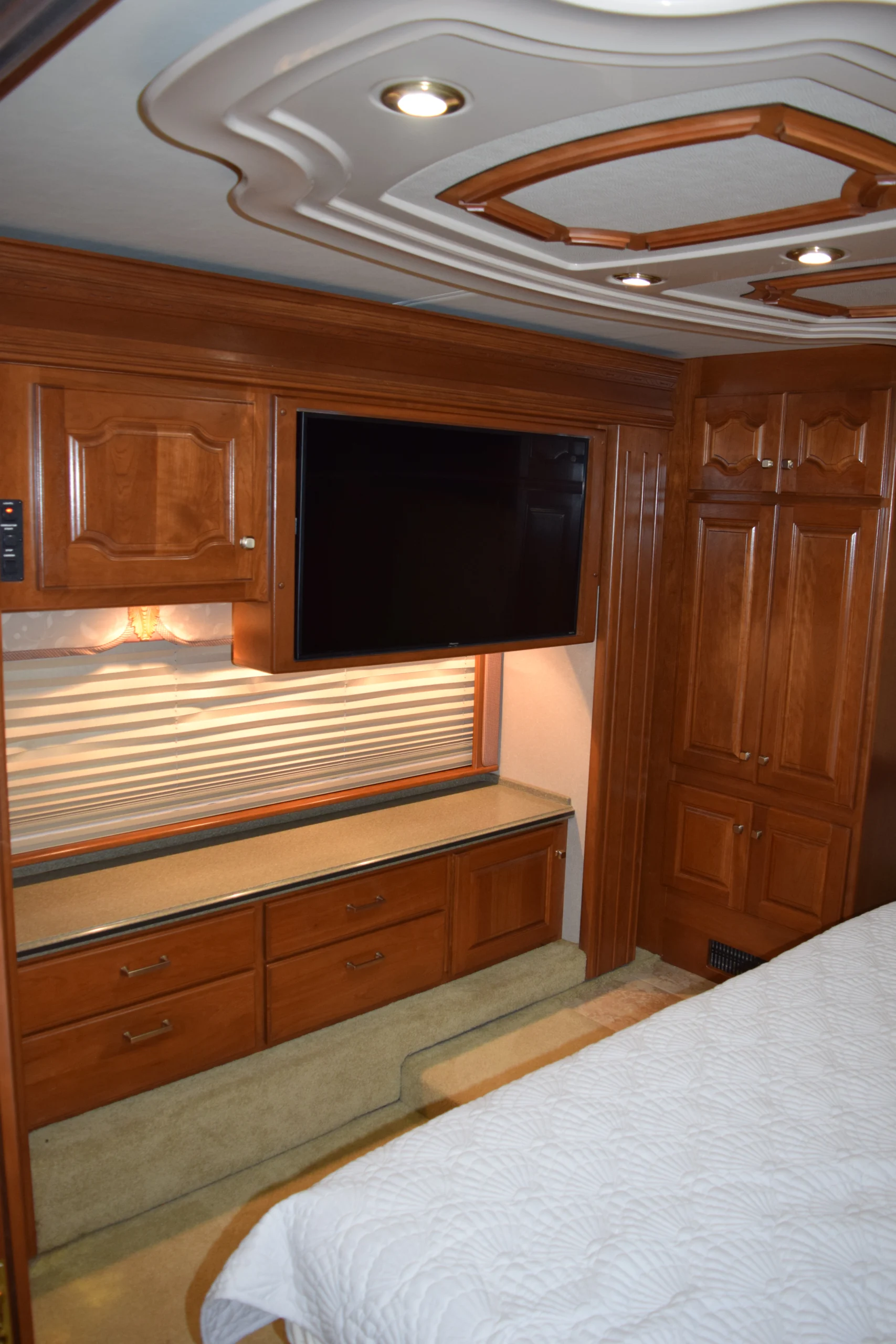 DSC_0193 Luxurious Motorcoach and RVs interior with wooden cabinets, mounted TV, decorative ceiling, and a bed with white quilt.
