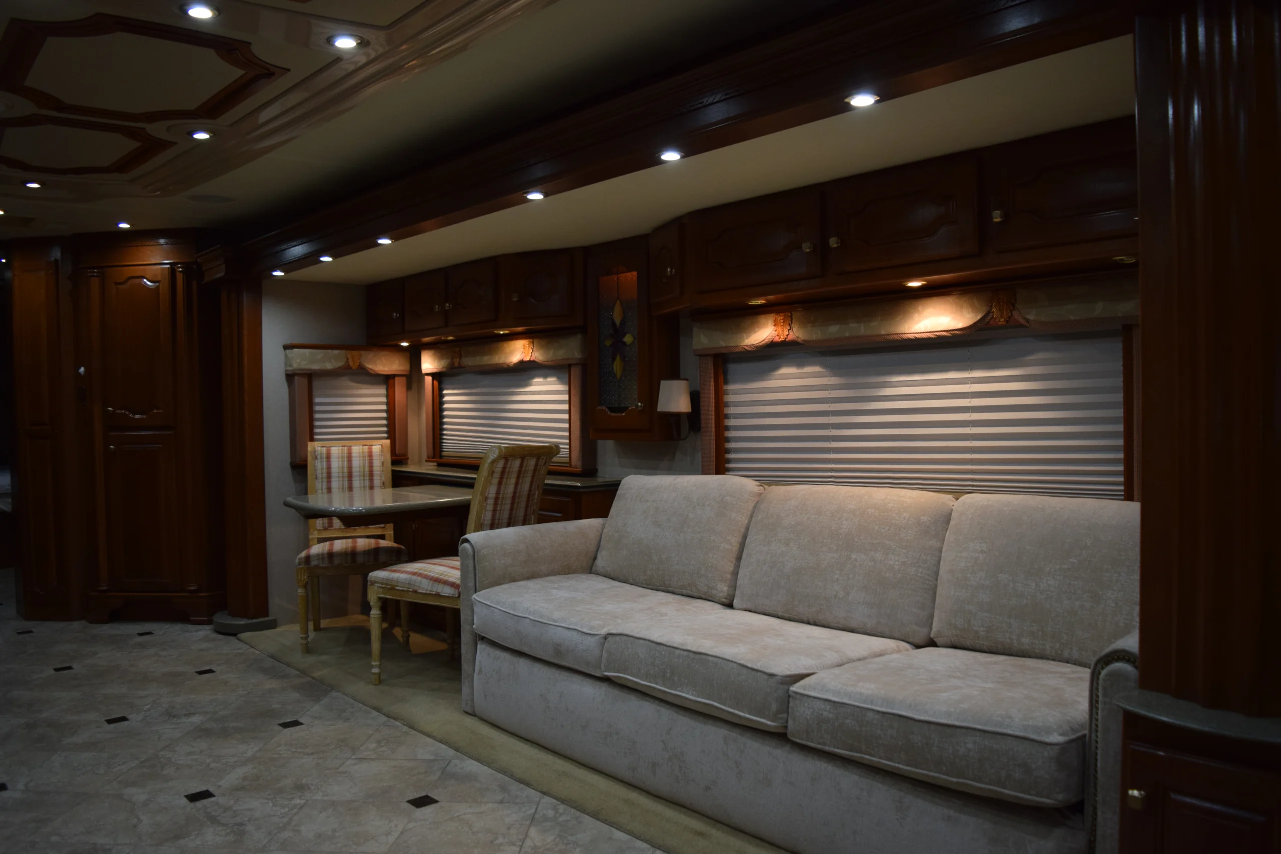 DSC_0210 Luxurious motorcoach interior featuring a beige sofa, dining area, elegant wood cabinetry, and soft ambient lighting.