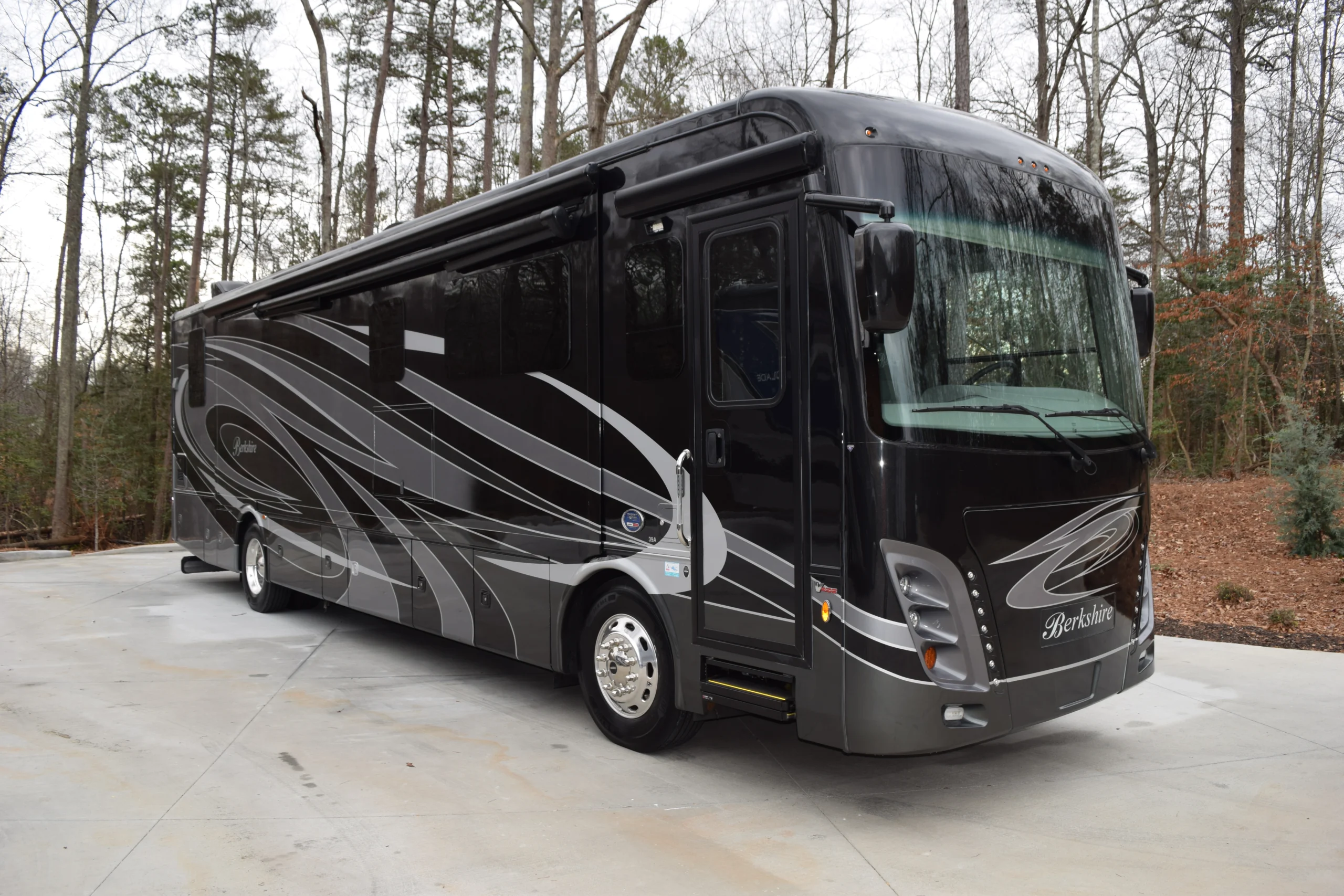 DSC_0240 Luxury black motorcoach RV with silver swirls parked on a concrete surface, surrounded by tall trees.