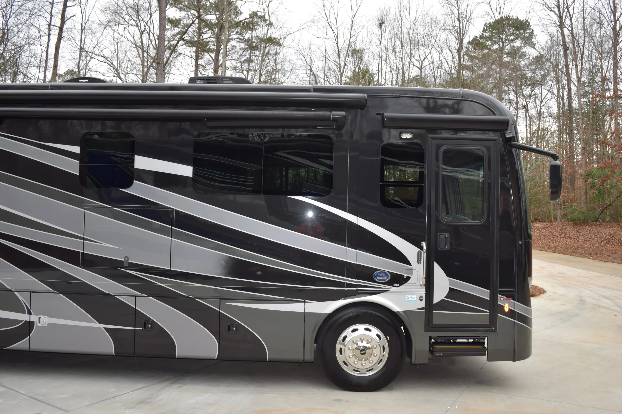 DSC_0242 Black motorcoach with sleek gray and white stripes, parked on a concrete lot surrounded by bare trees.