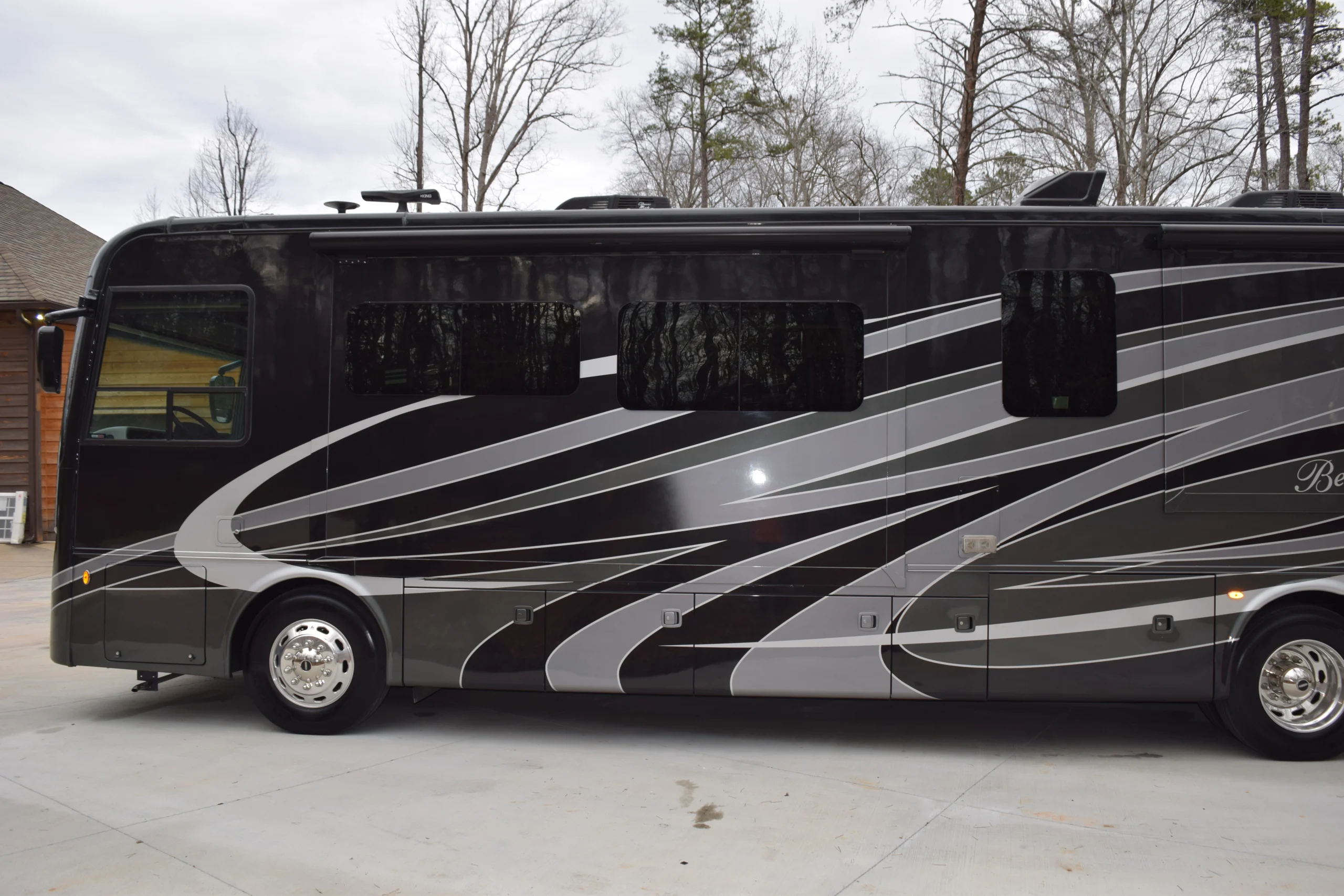 DSC_0250 Sleek black motorcoach with silver and white stripes parked on a driveway beside a wooden building, surrounded by bare trees.