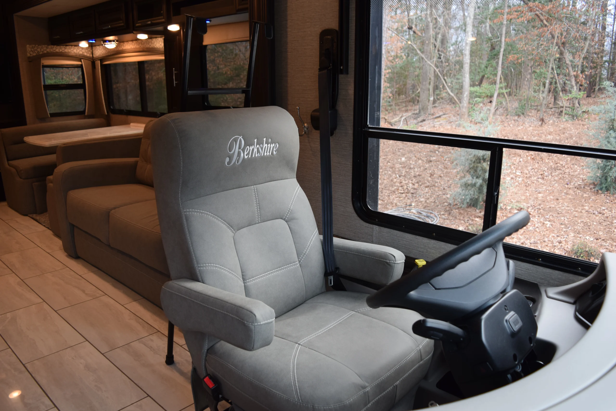 DSC_0292 Driving seat and dashboard of a luxury motorcoach, with plush seating, polished flooring, and scenic forest view through large windows.