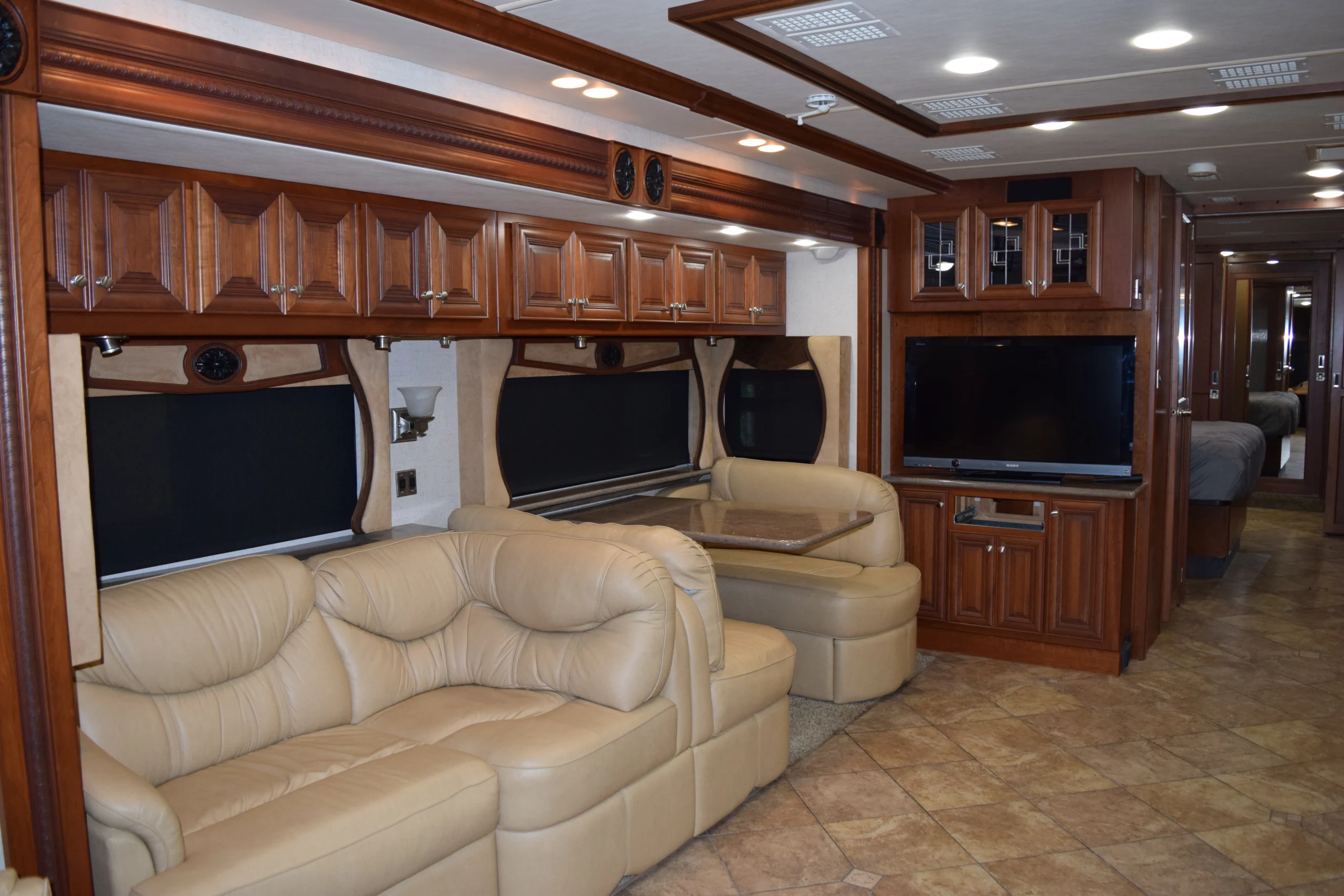 DSC_0333 Luxurious Motorcoach interior with leather seating, wood cabinets, a large TV, and a cozy dinette area leading to a bedroom.