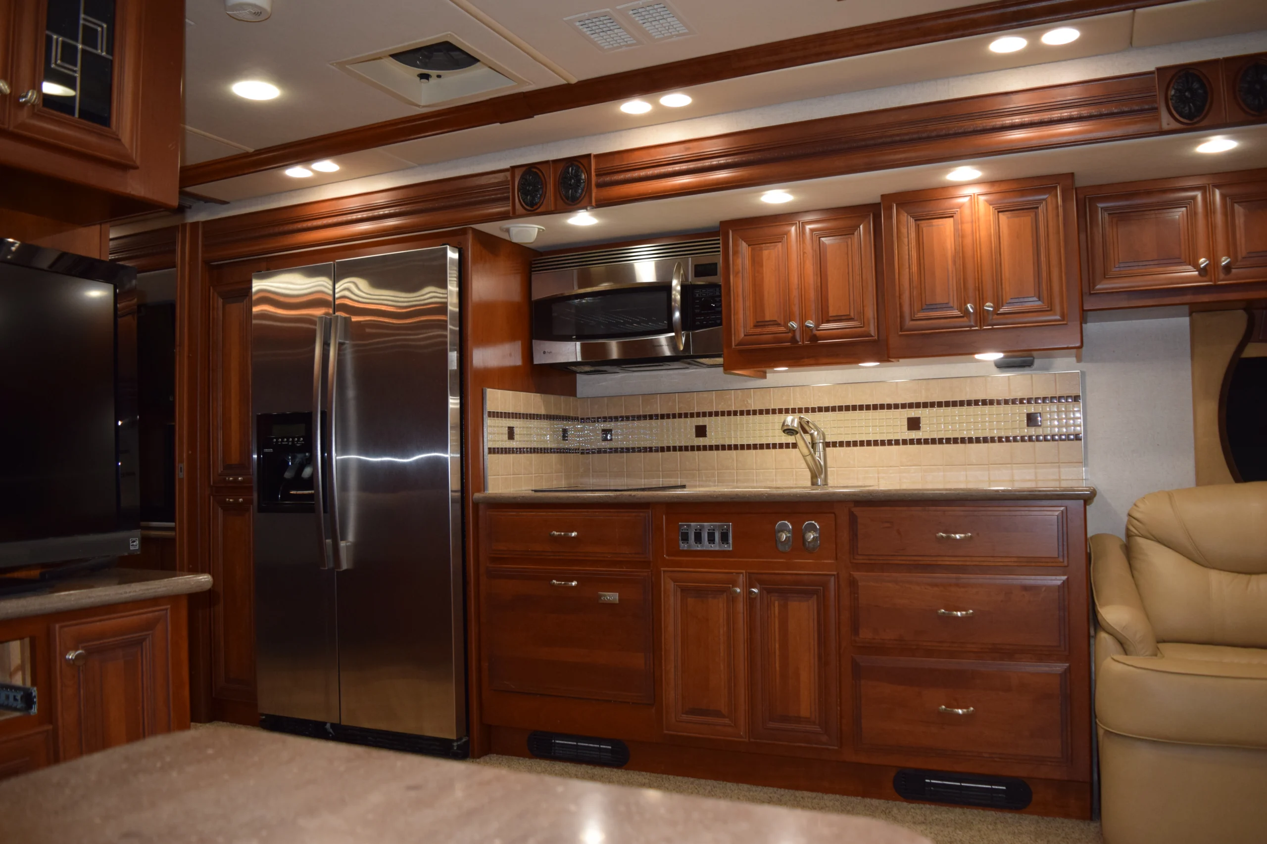 DSC_0345 Spacious motorcoach kitchen with wooden cabinets, stainless steel fridge, microwave, and beige tile backsplash, adjacent to leather seating.