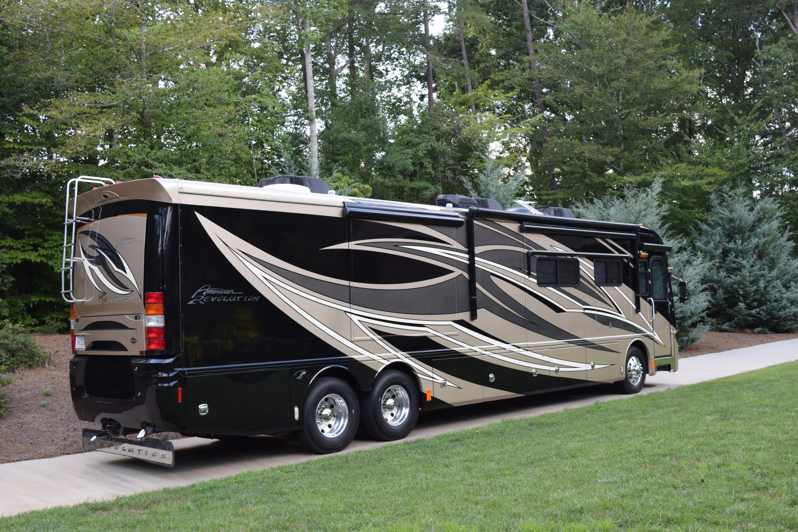 DSC_0346 Luxury motorcoach with sleek tan and black design parked on driveway, surrounded by lush green trees.