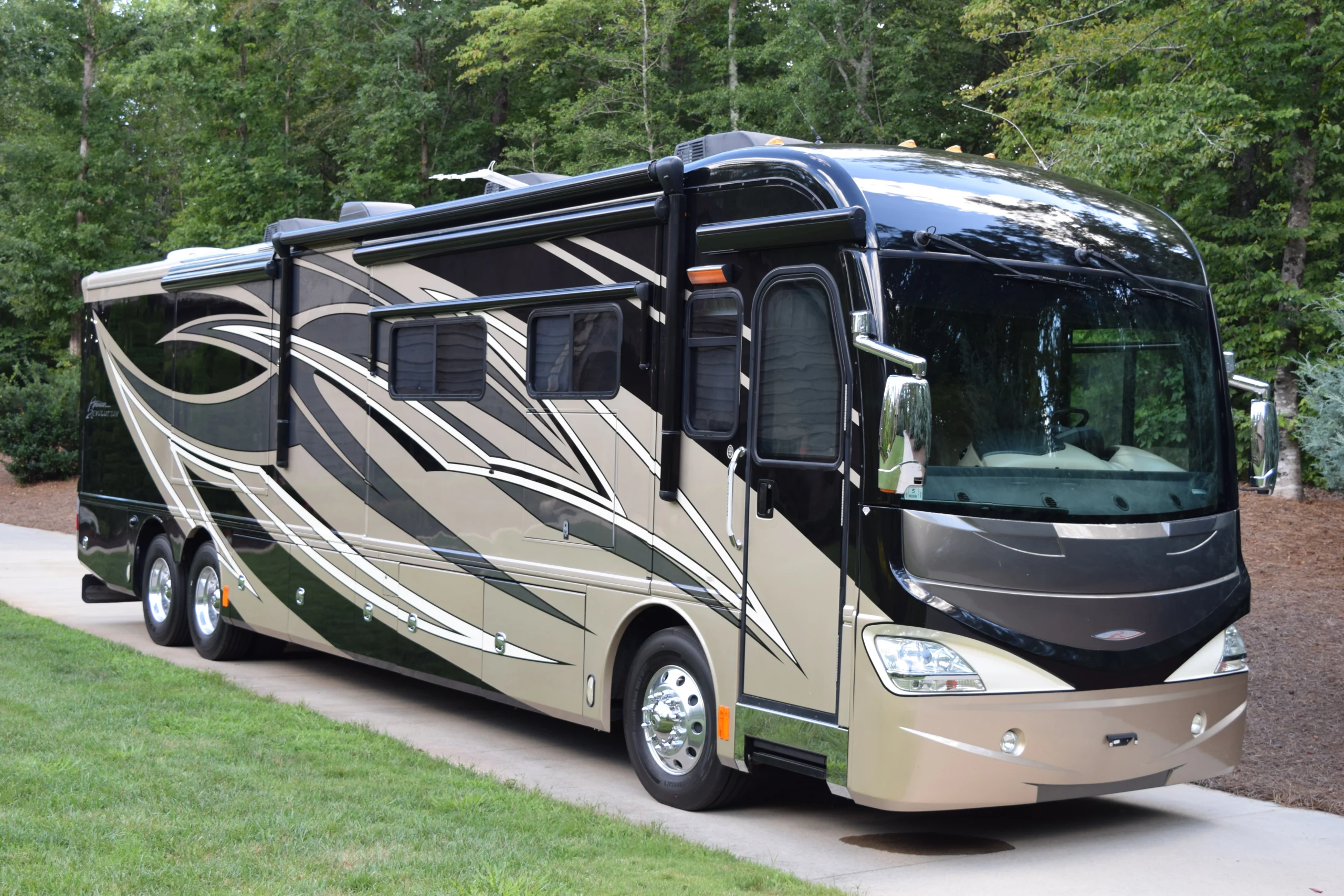 DSC_0347 Luxurious motorcoach parked on a driveway, featuring sleek, modern design with dark and beige swirls, set against a lush green backdrop.