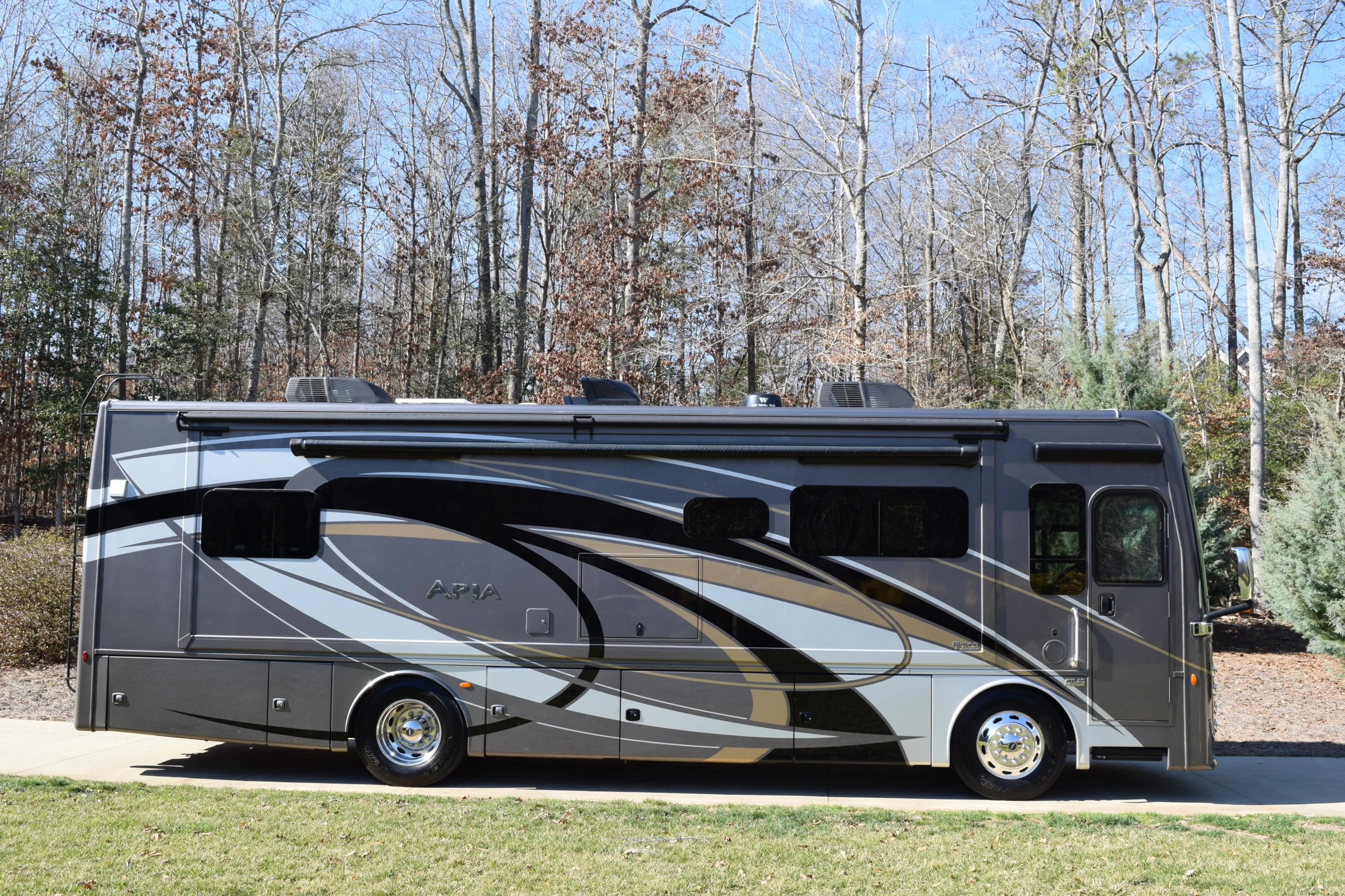 DSC_0372 Sleek motorcoach RV parked on driveway surrounded by bare trees, featuring a stylish silver and black design.