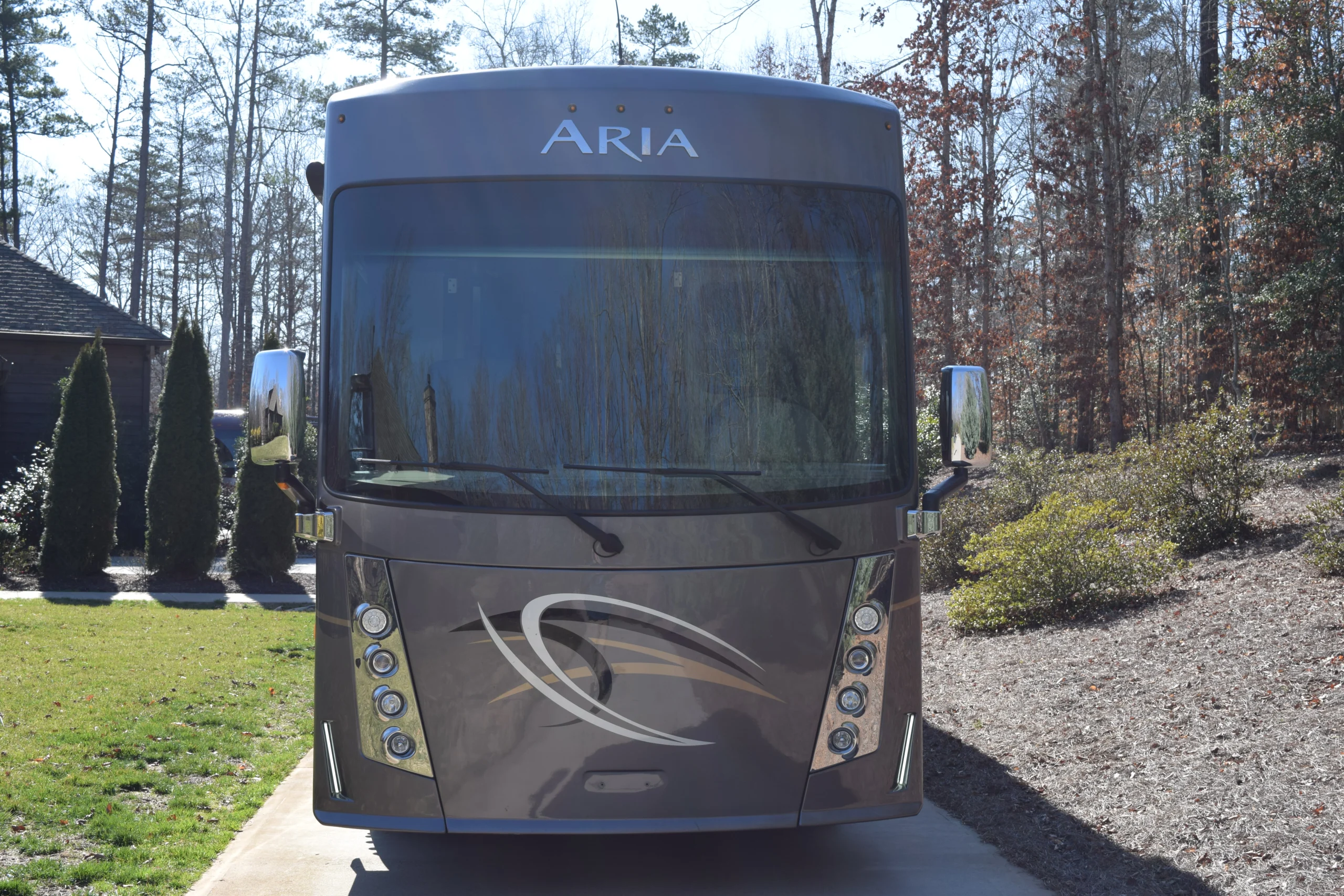 DSC_0374 Front view of a large, sleek motorcoach named Aria parked on a driveway, surrounded by trees and bushes.