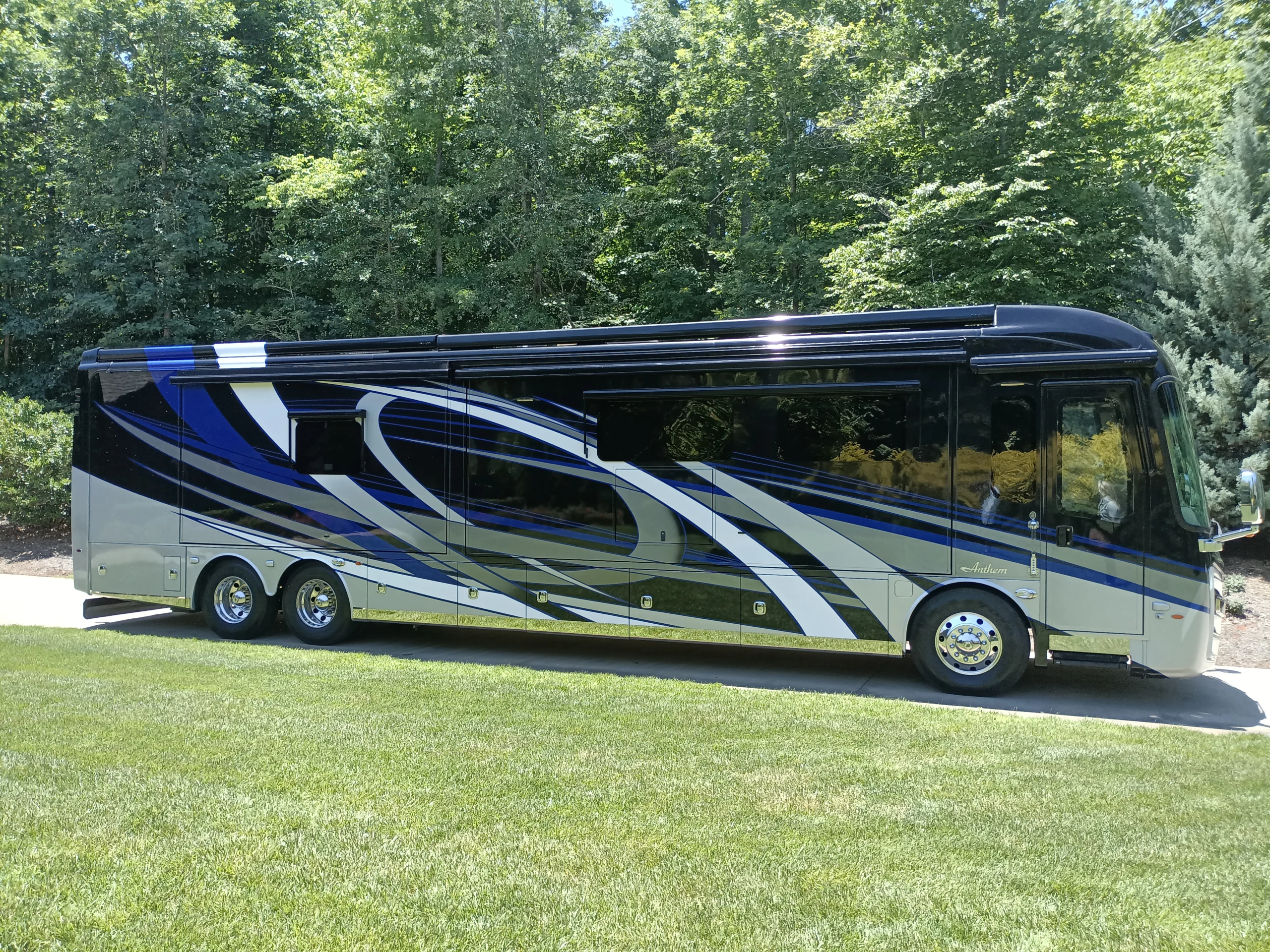 IMG_20260603_131837_452 Luxurious motorcoach parked on a grassy lawn with blue, white, and silver details, surrounded by lush green trees.