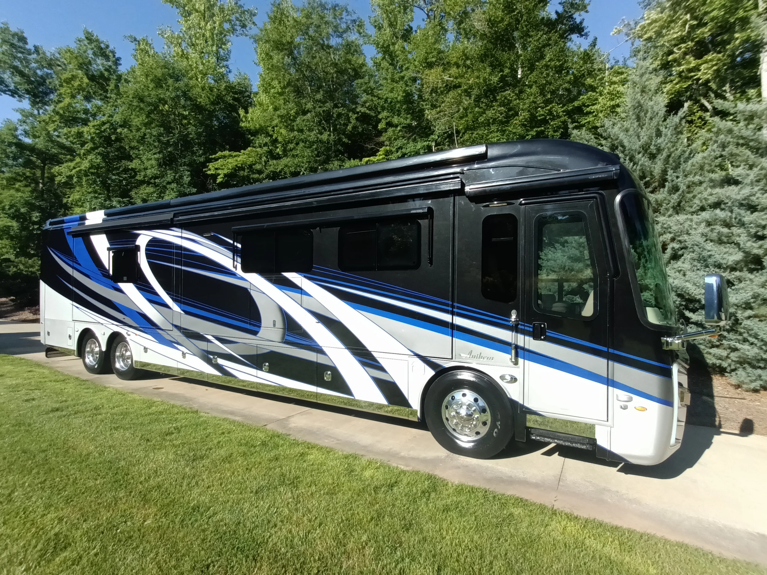 IMG_20260604_084156_835 Luxury Motorcoach with blue and white swirls parked on a driveway, surrounded by lush green trees.