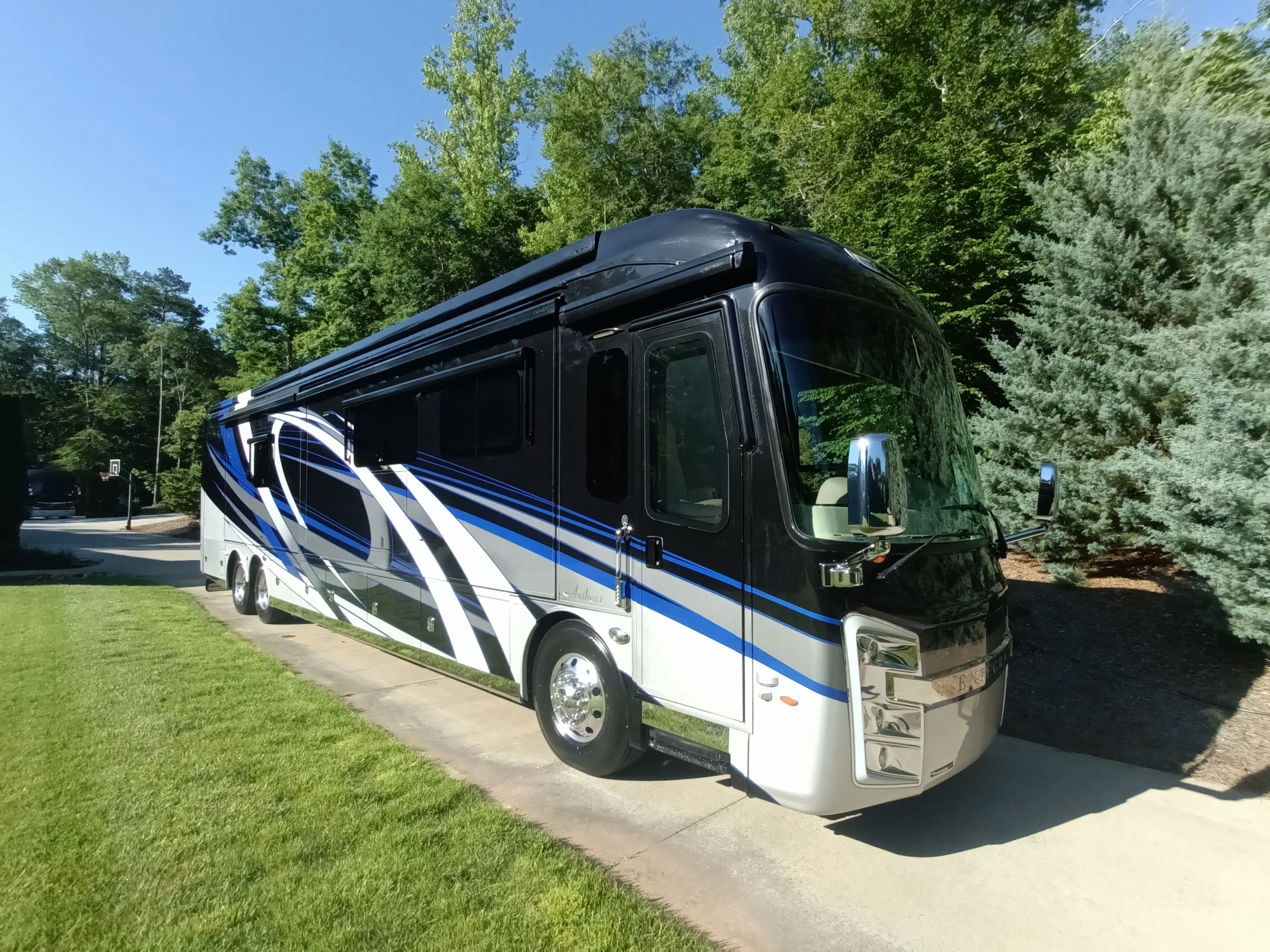 IMG_20260604_084203_855 Sleek black and blue motorcoach parked on a sunny driveway, surrounded by lush green trees.