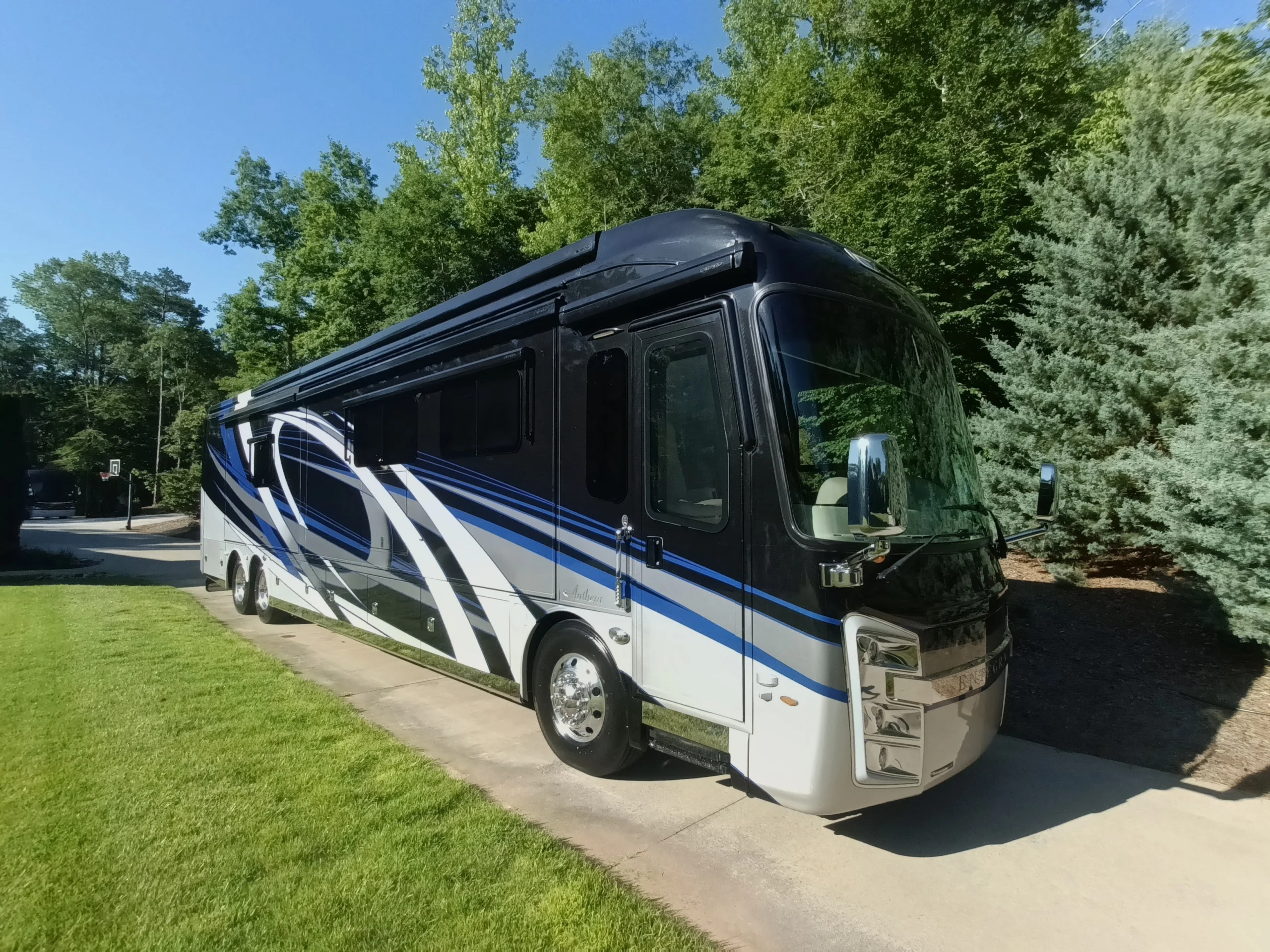 IMG_20260604_084203_855~2 Luxury motorcoach parked on driveway; sleek black and blue design. Surrounded by lush greenery and a clear blue sky.