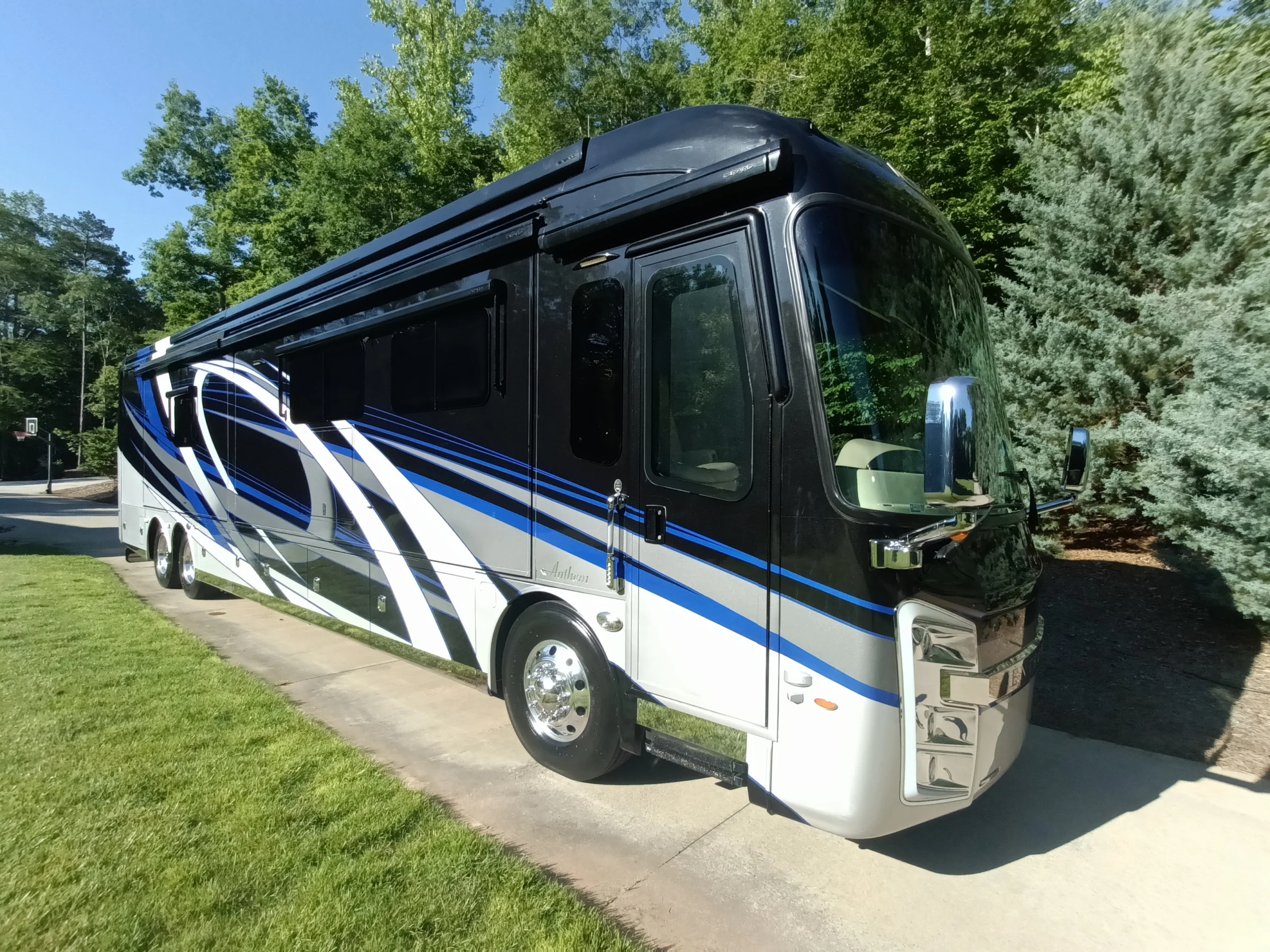 IMG_20260604_084208_893 Sleek black motorcoach with blue and white accents parked on a driveway, surrounded by lush greenery and trees.