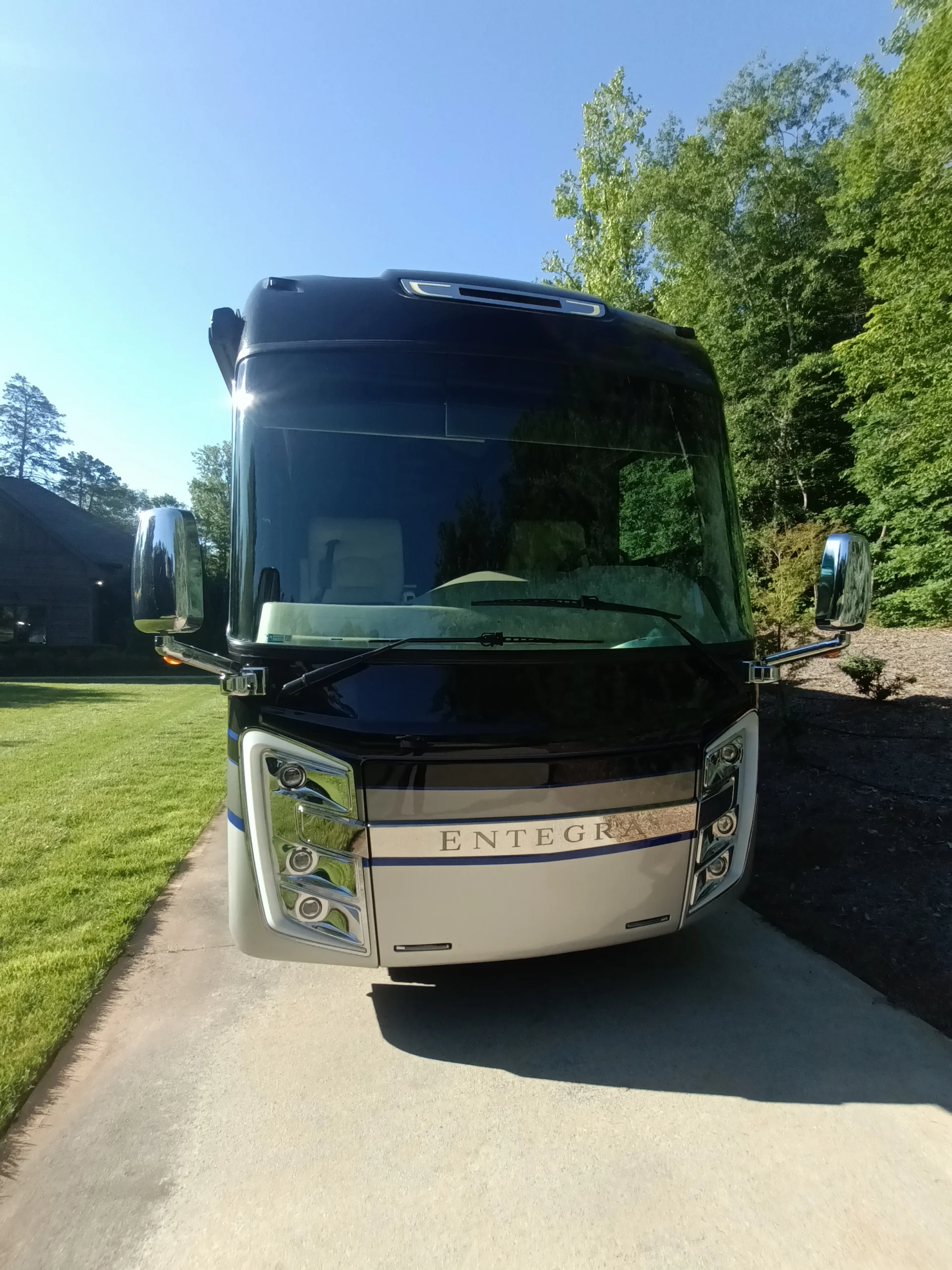 IMG_20260604_084221_061 Front view of a luxury Motorcoach under clear sky, parked on a driveway with lush greenery in the background.