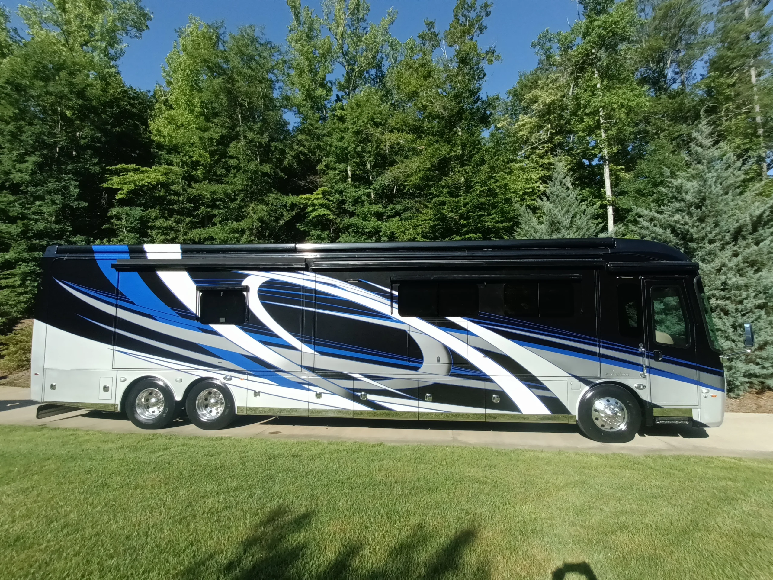 IMG_20260604_084255_263 Motorcoach with blue, black, and white swirl design parked on grass, surrounded by dense green trees under a clear blue sky.