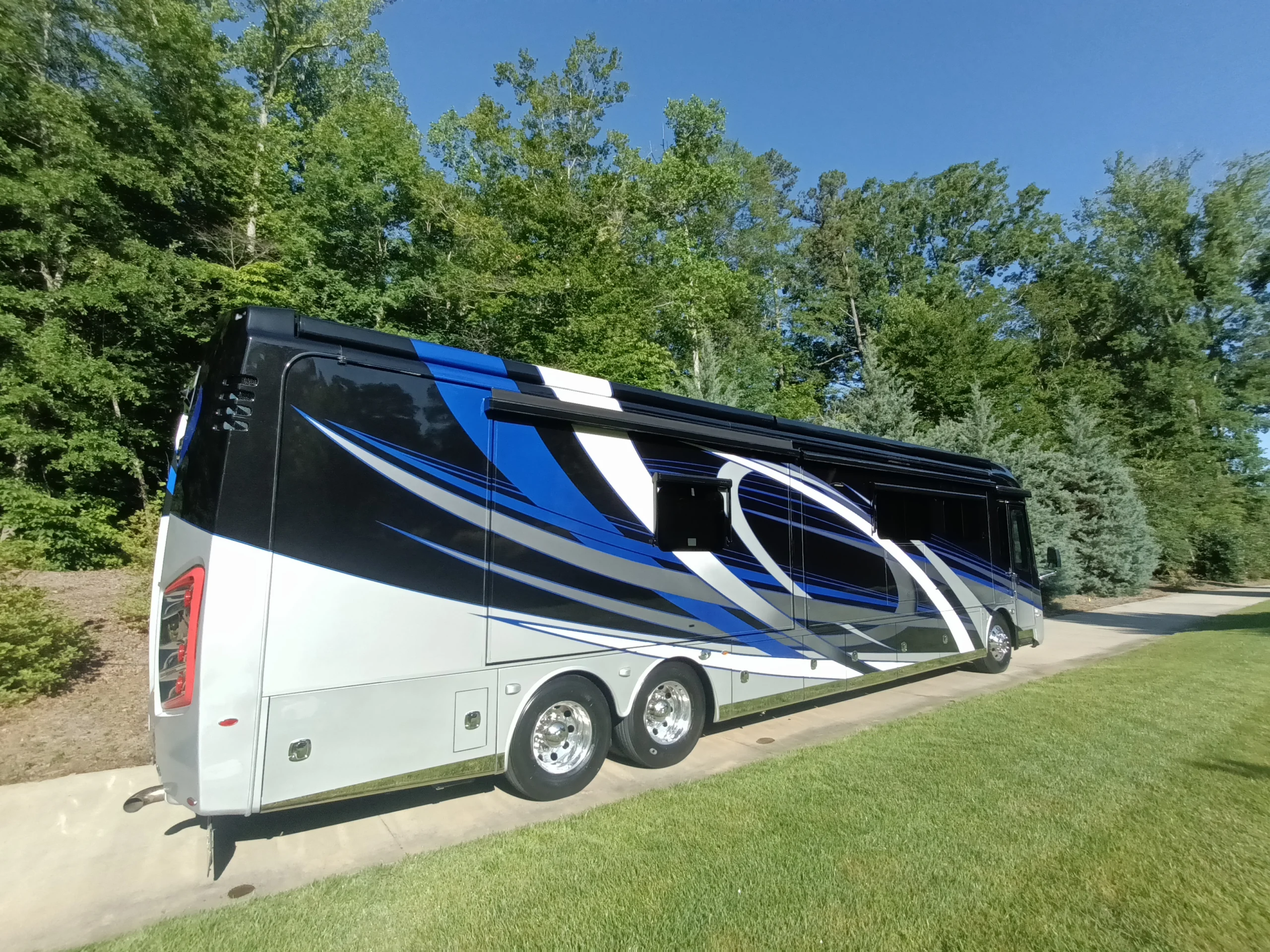 IMG_20260604_084311_754 Large motorcoach parked on a sunny driveway, featuring blue, white, and black swirling designs, surrounded by green trees and grass.