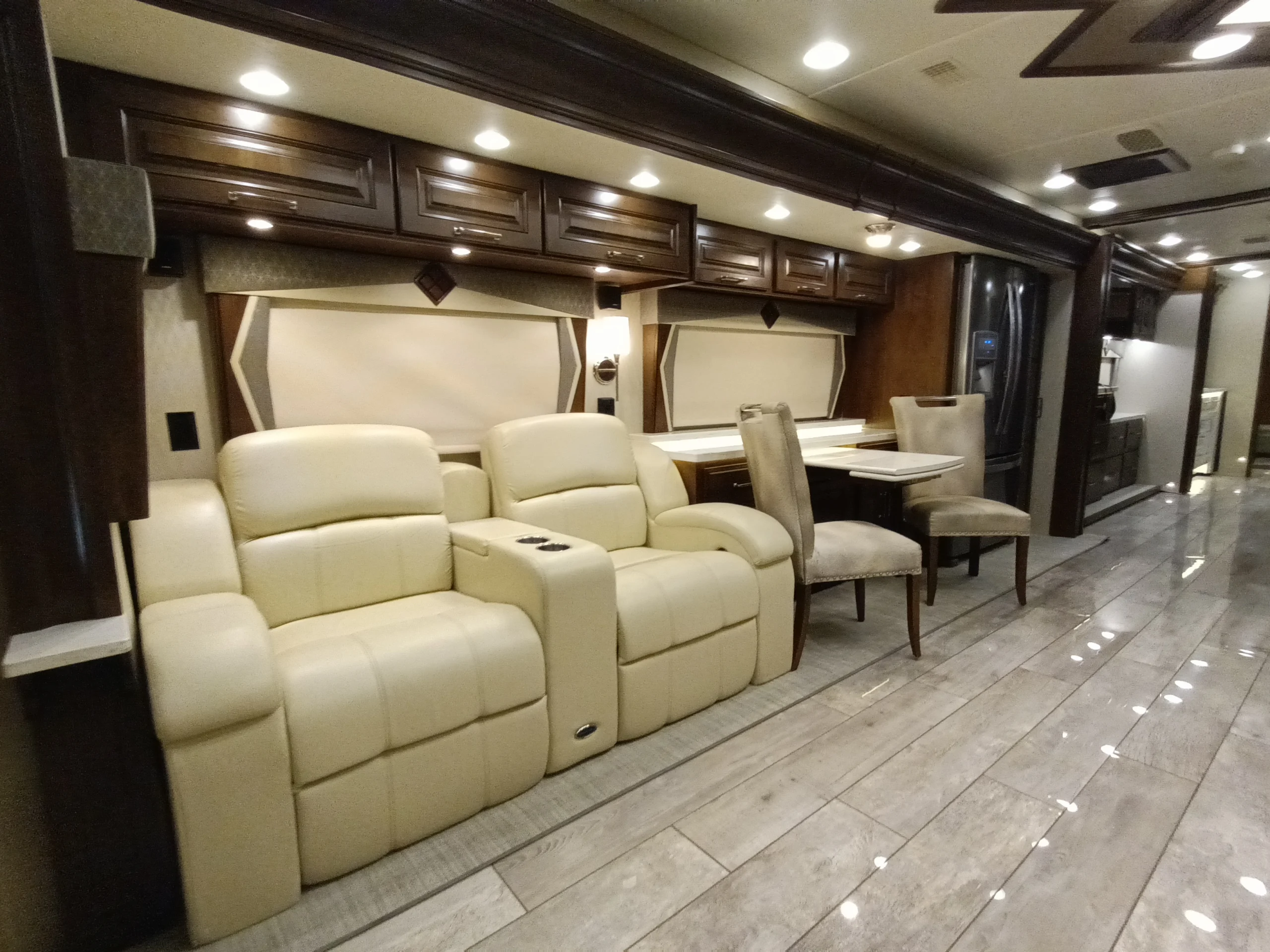 IMG_20260604_122300_122 Luxurious Motorcoach interior with cream leather recliners, plush dining chairs, elegant wood cabinetry, and glossy tile flooring.