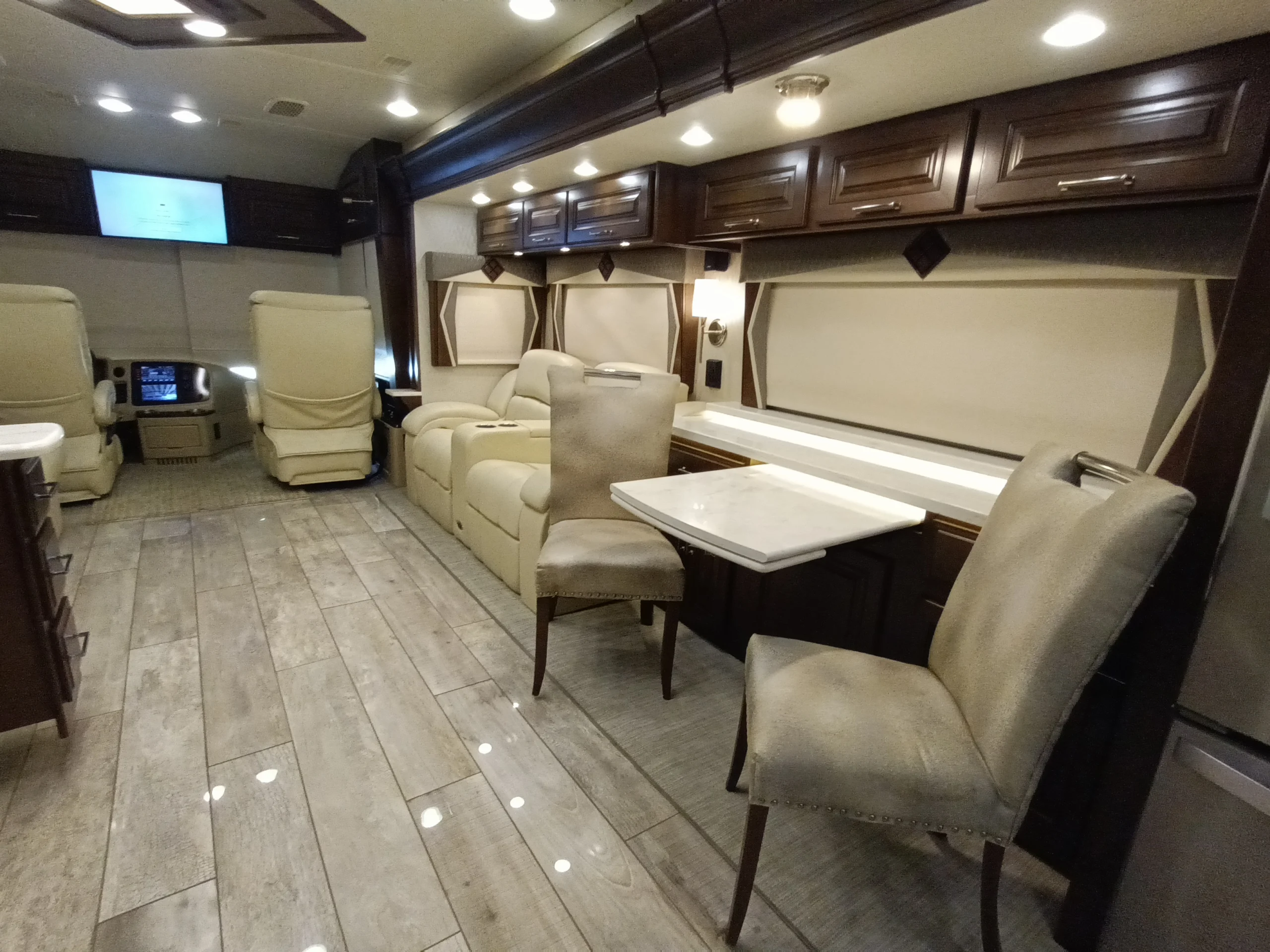 IMG_20260604_122313_793 Spacious Motorcoach interior with beige leather seating, dining area, flat-screen TV, and wood-like flooring under warm recessed lighting.